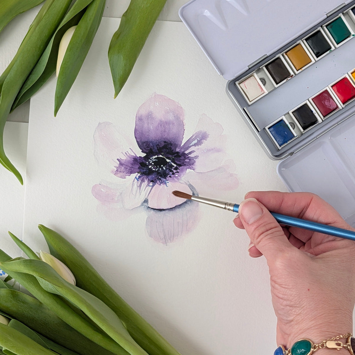 Person painting a purple flower with watercolors on a white surface, surrounded by green leaves.