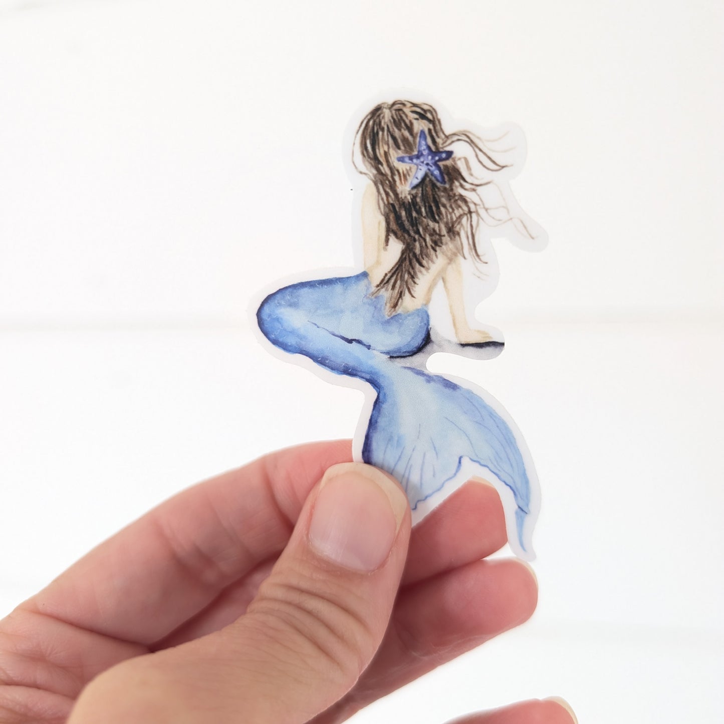 Hand holding a sticker of a mermaid with a blue tail and starfish.
