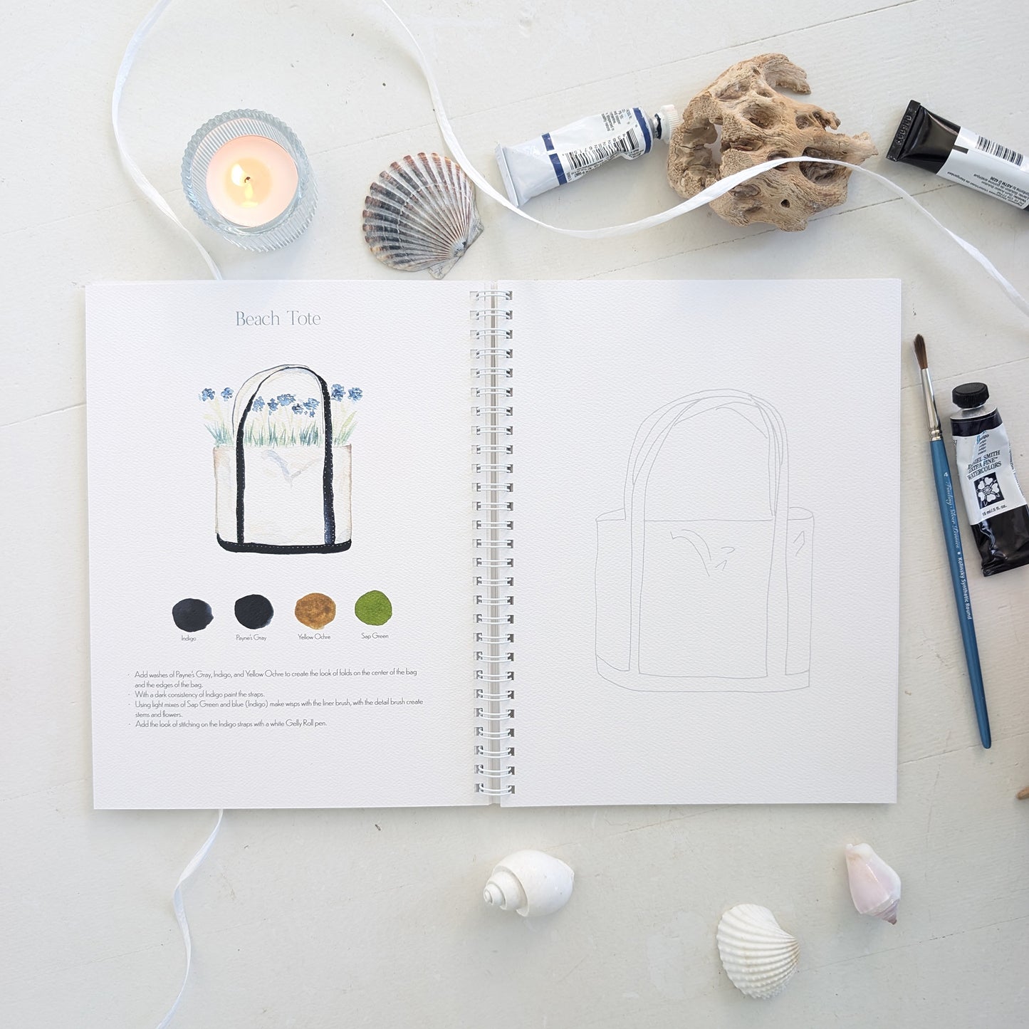 Open watercolor workbook with beach tote illustrations on a surface with art supplies and shells