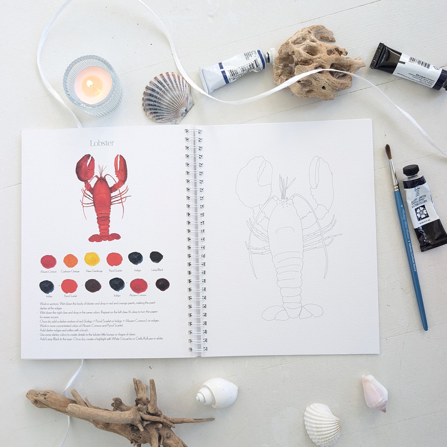 Open watercolor workbook with a lobster illustration and color swatches, surrounded by art supplies on a light surface.