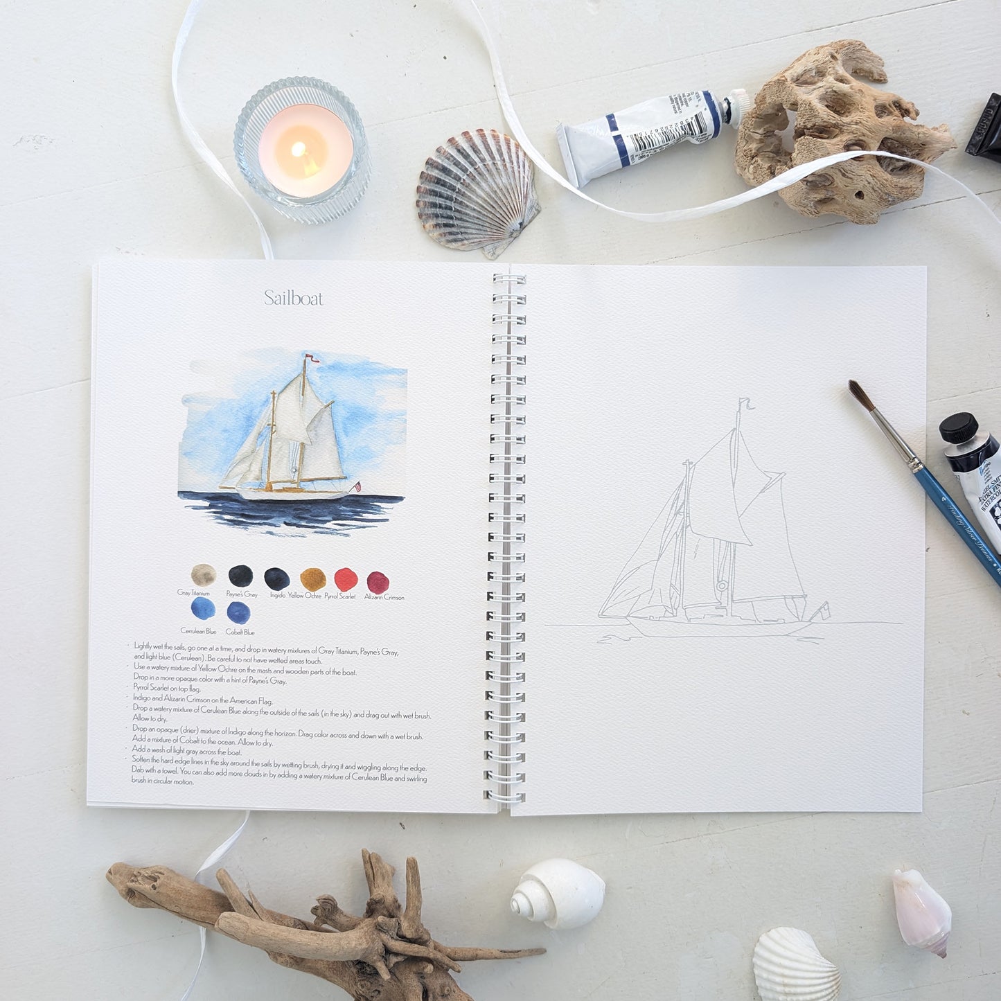Open watercolor workbook with sailboat illustration and color swatches on a white surface with natural elements.