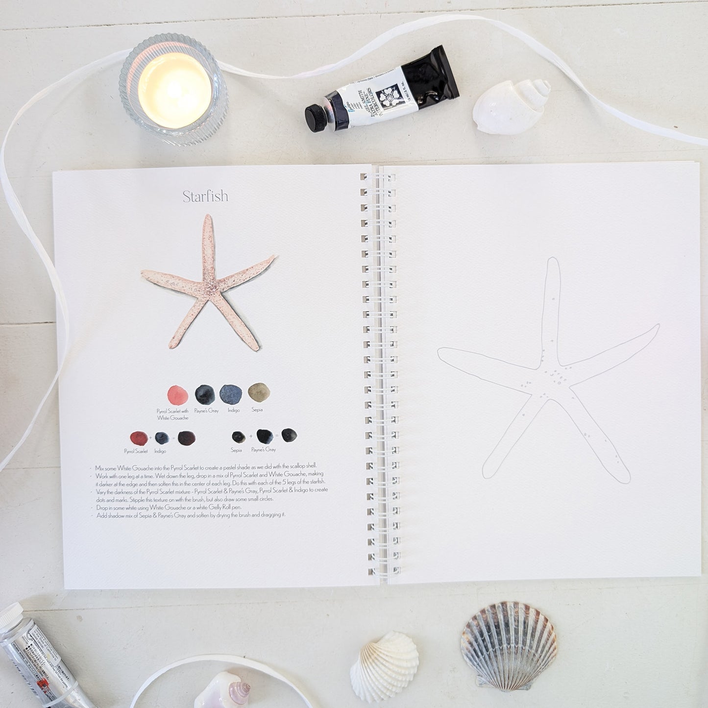 Open watercolor workbook with starfish illustration and color swatches on a white surface with a candle and shells.