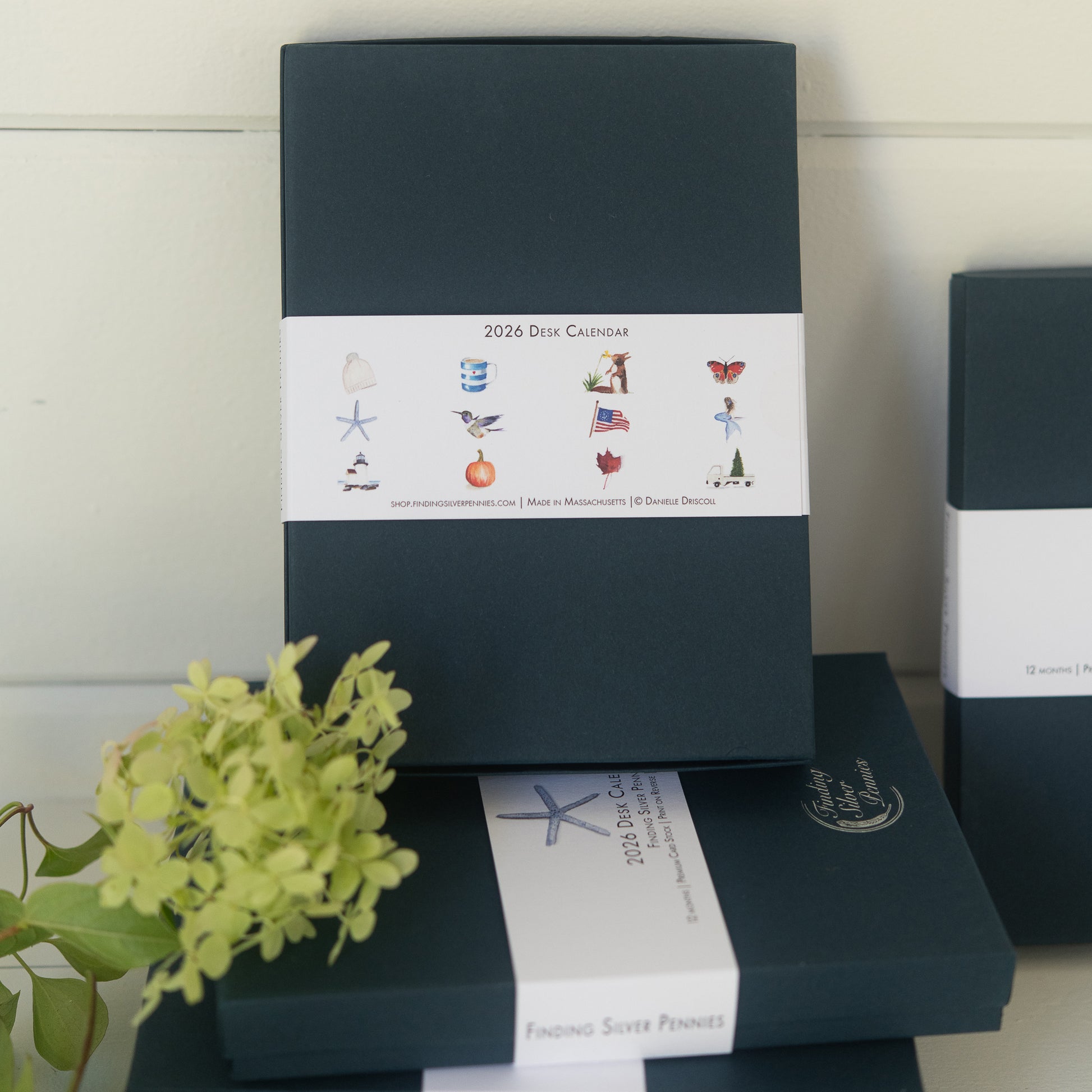 Stack of dark blue boxes with white labels featuring watercolor illustrations and text on a light background. 2026 Desk Calendar boxes.
