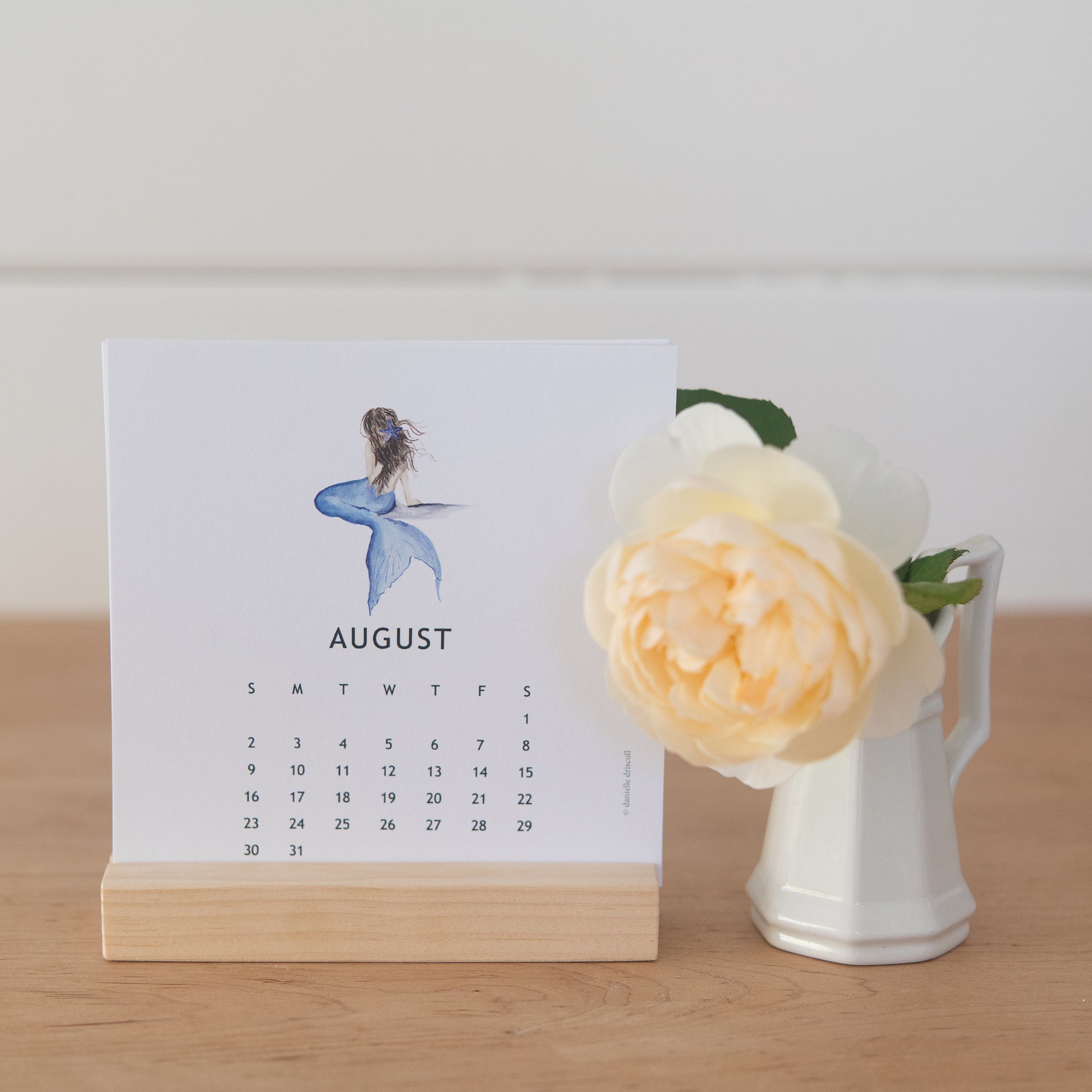Calendar for August with a mermaid design in a wooden stand next to a white vase with a blush rose.