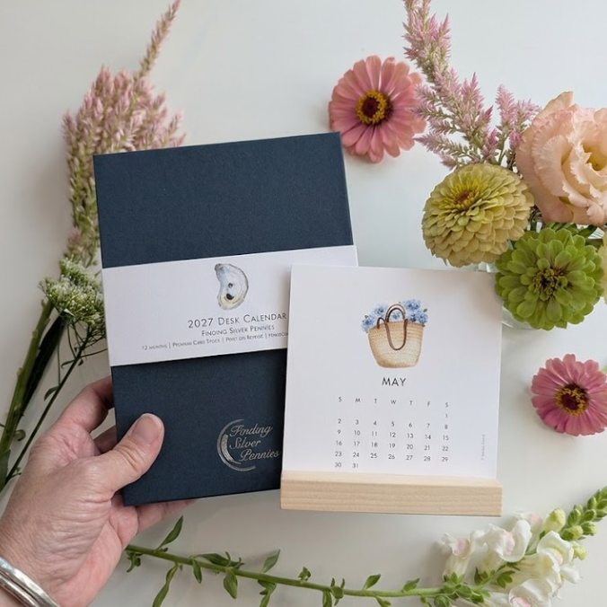 A hand holds a navy blue 2027 Year Calendar box next to an open May calendar page on a wooden stand, styled with pink and green flowers on a white background.