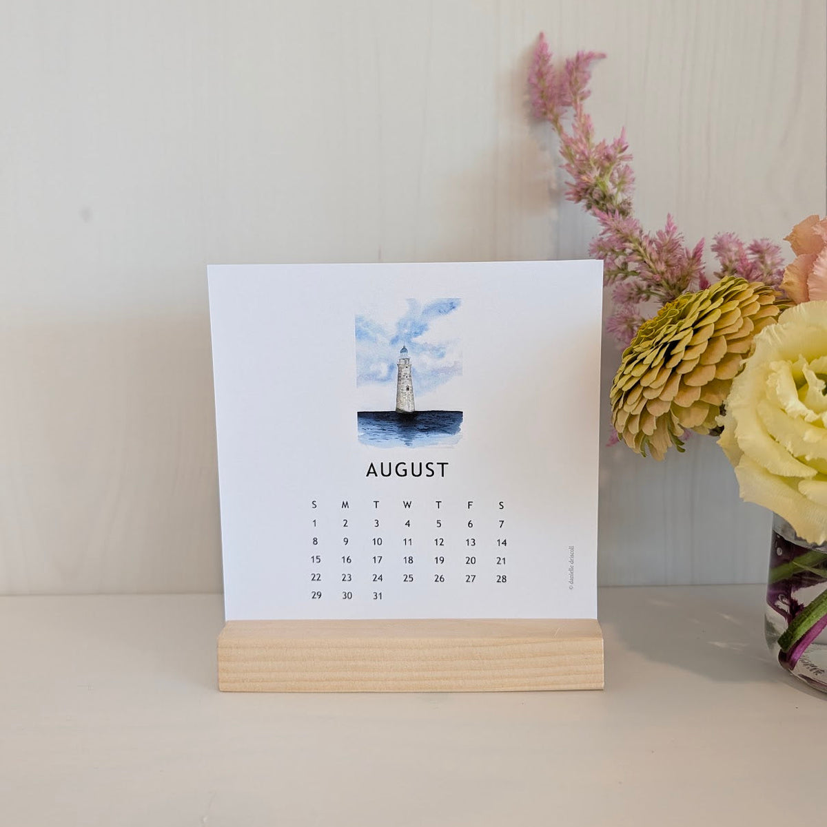 A single-page desk calendar standing on a light wood block, showing August with a watercolor lighthouse illustration. The calendar is photographed on a tabletop against a pale wall, with flowers partially visible on the right.