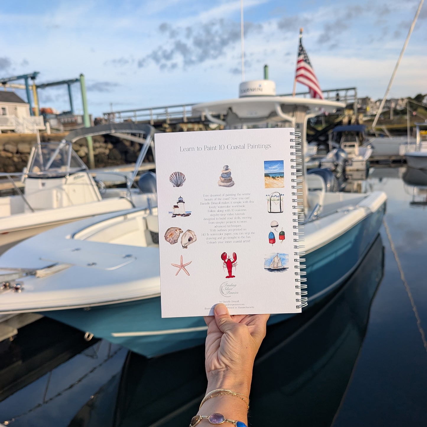 Person holding a watercolor workbook with illustrations of coastal elements in front of boats and a dock.