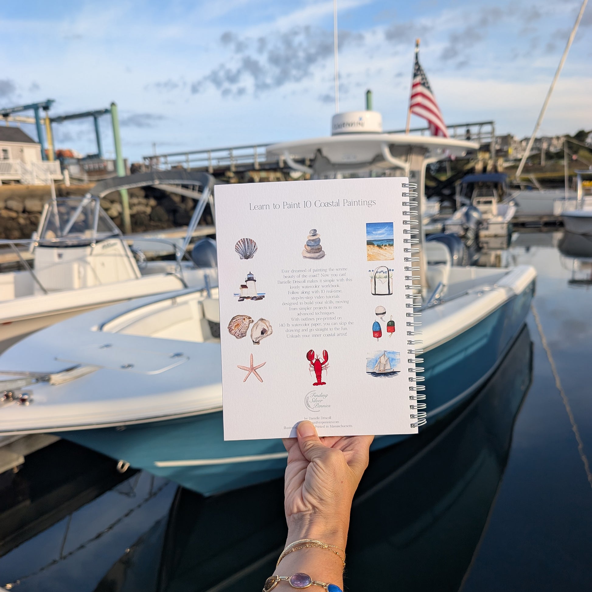 Person holding a watercolor workbook with illustrations of coastal elements in front of boats and a dock.