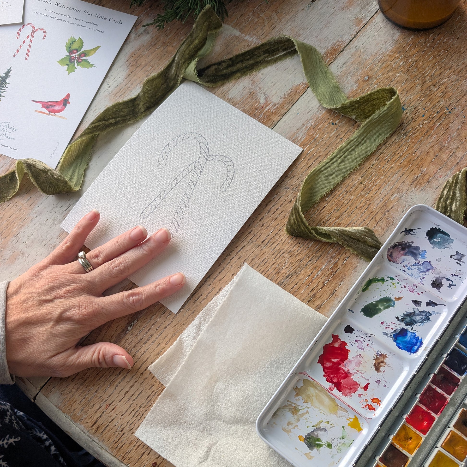 Hand holding a piece of paper with a candy cane drawing on a wooden table with watercolor supplies.