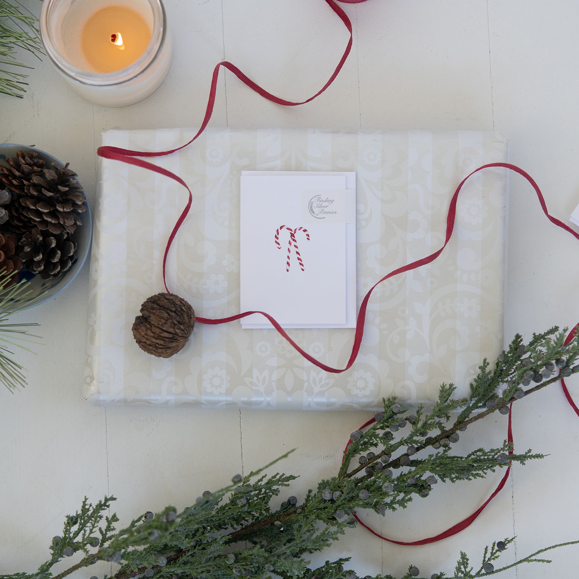 Gift wrapped in white paper with a red ribbon, surrounded by greenery and a candle.