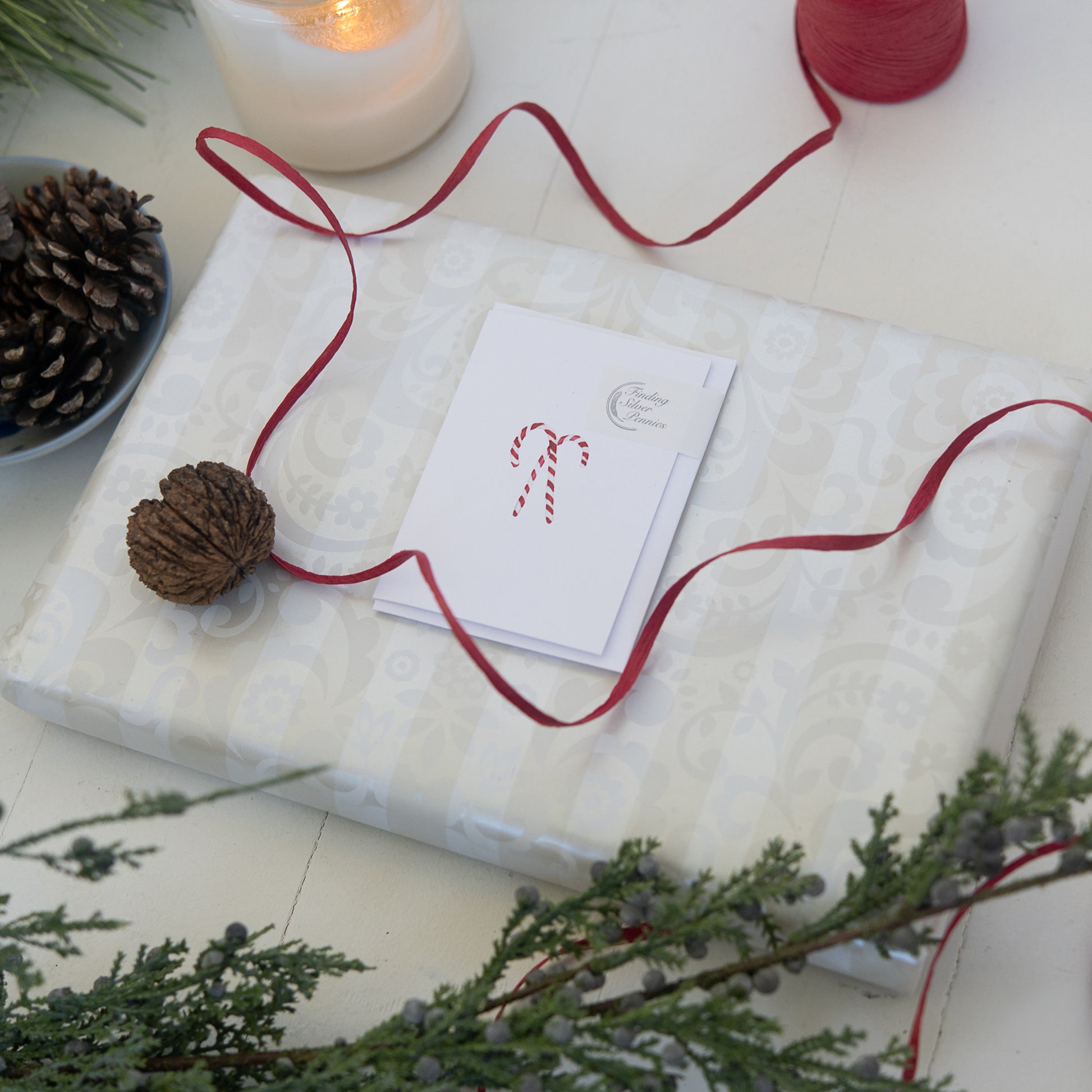 Gift wrapped in white paper with a red ribbon, card, and pine cone on a festive background.