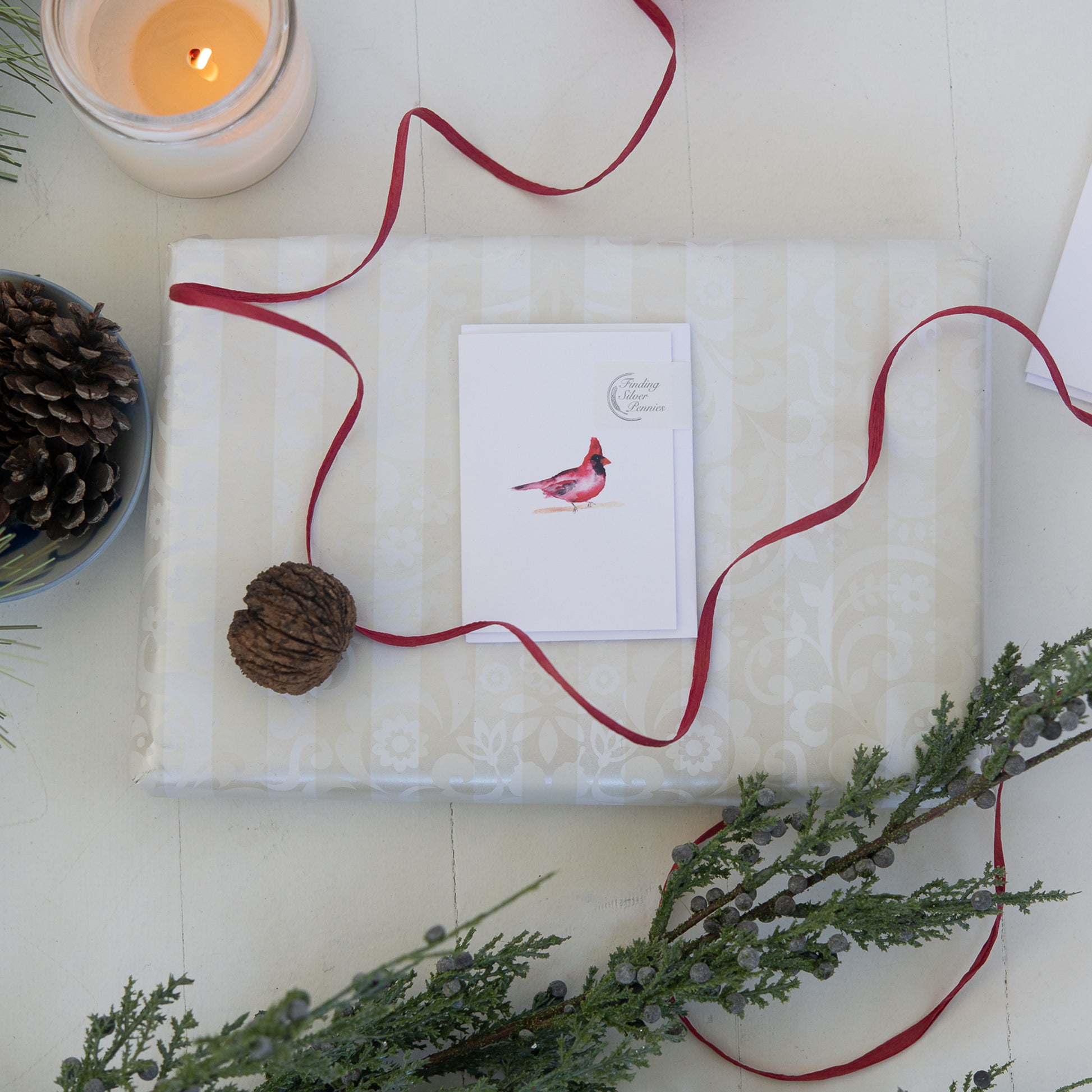 Gift wrapped in white paper with a red ribbon, card, and pine cone on a light surface.