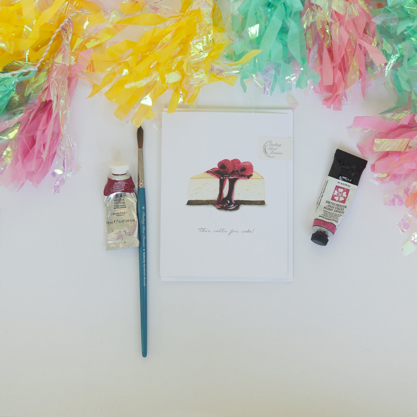 Artistic setup with a painting, paintbrush, and tubes of paint on a white surface with colorful tassels. cheesecake birthday card featured.