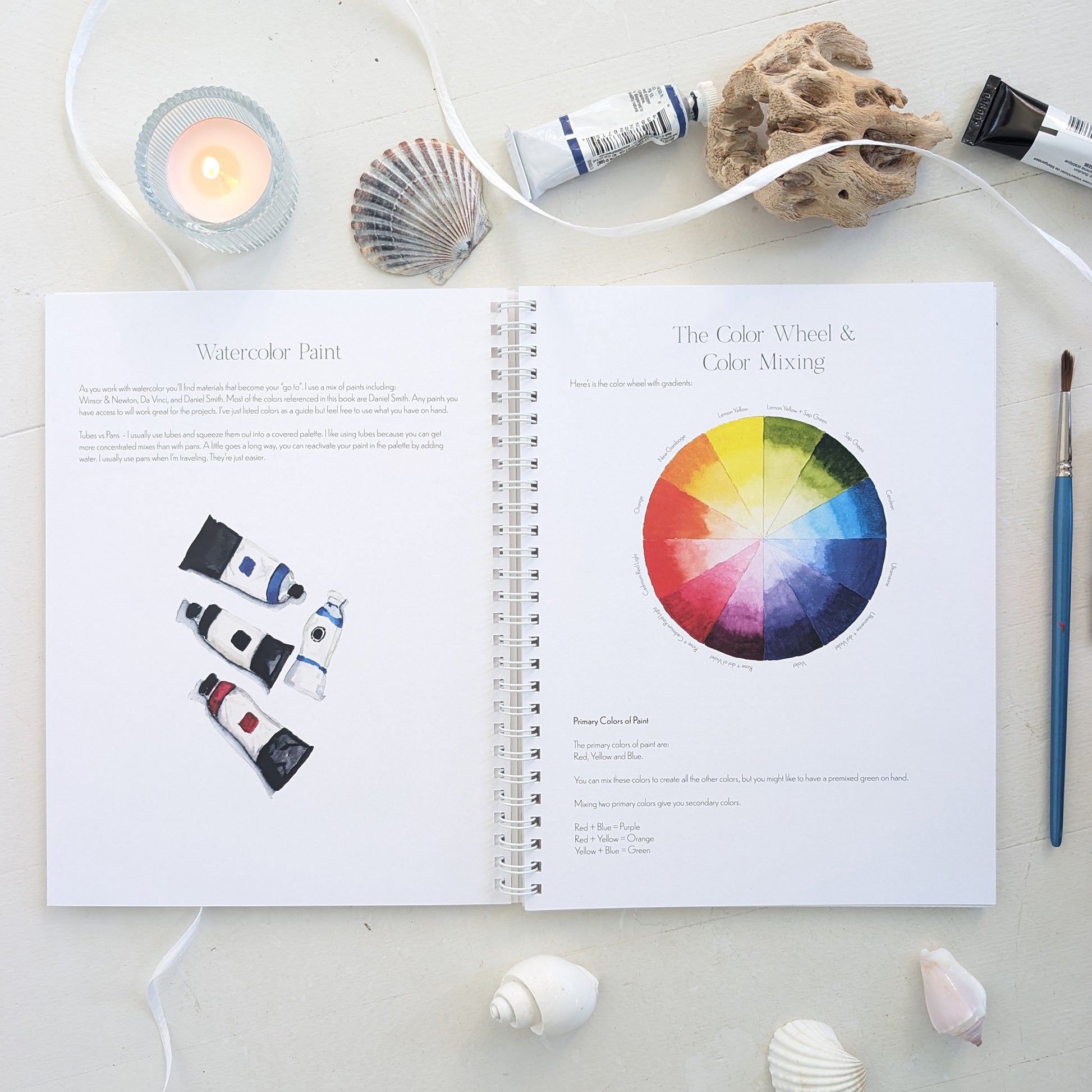 Open book on watercolor paint with a color wheel and art supplies on a white surface