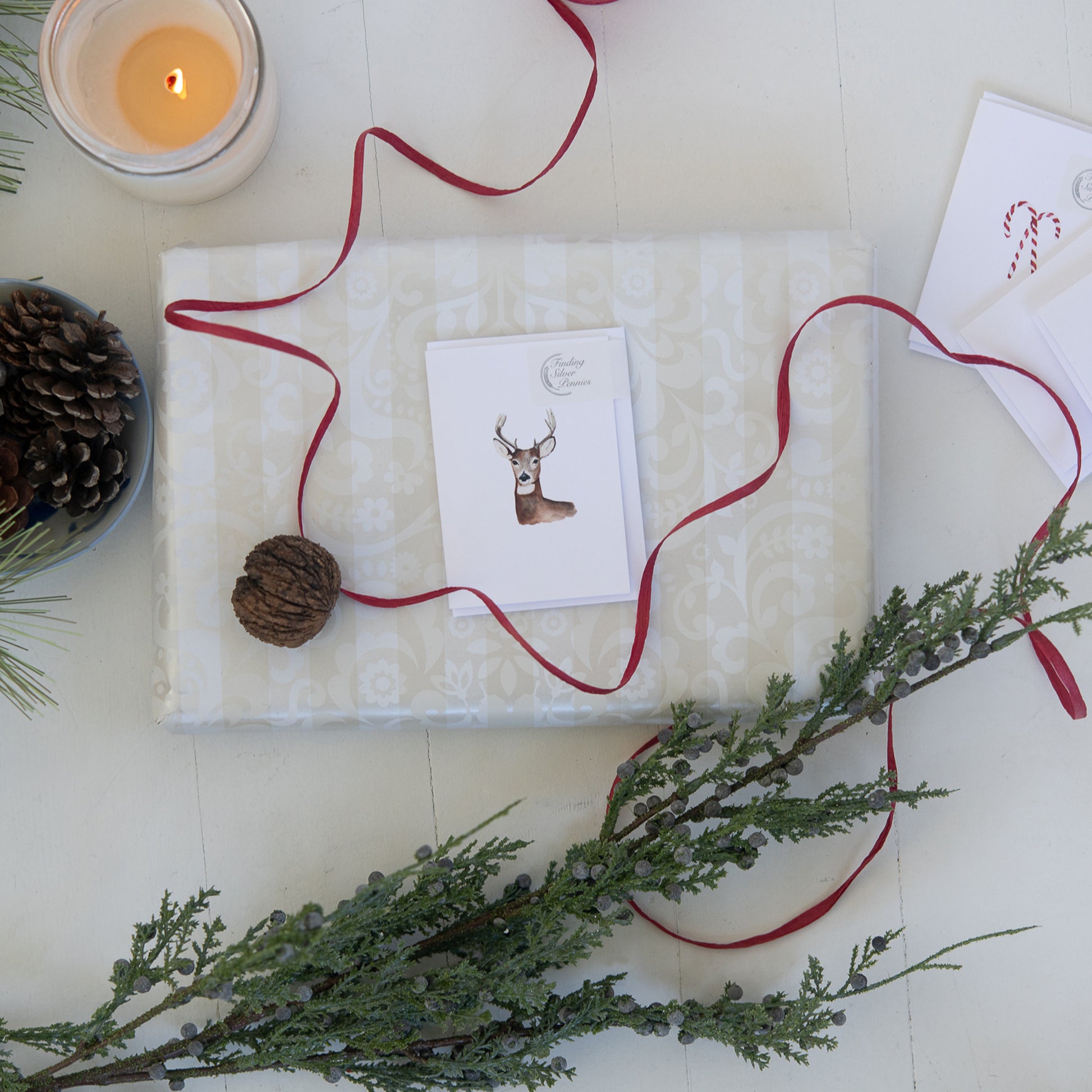 Gift wrapped in white paper with a red ribbon, card, and pinecones on a light surface.