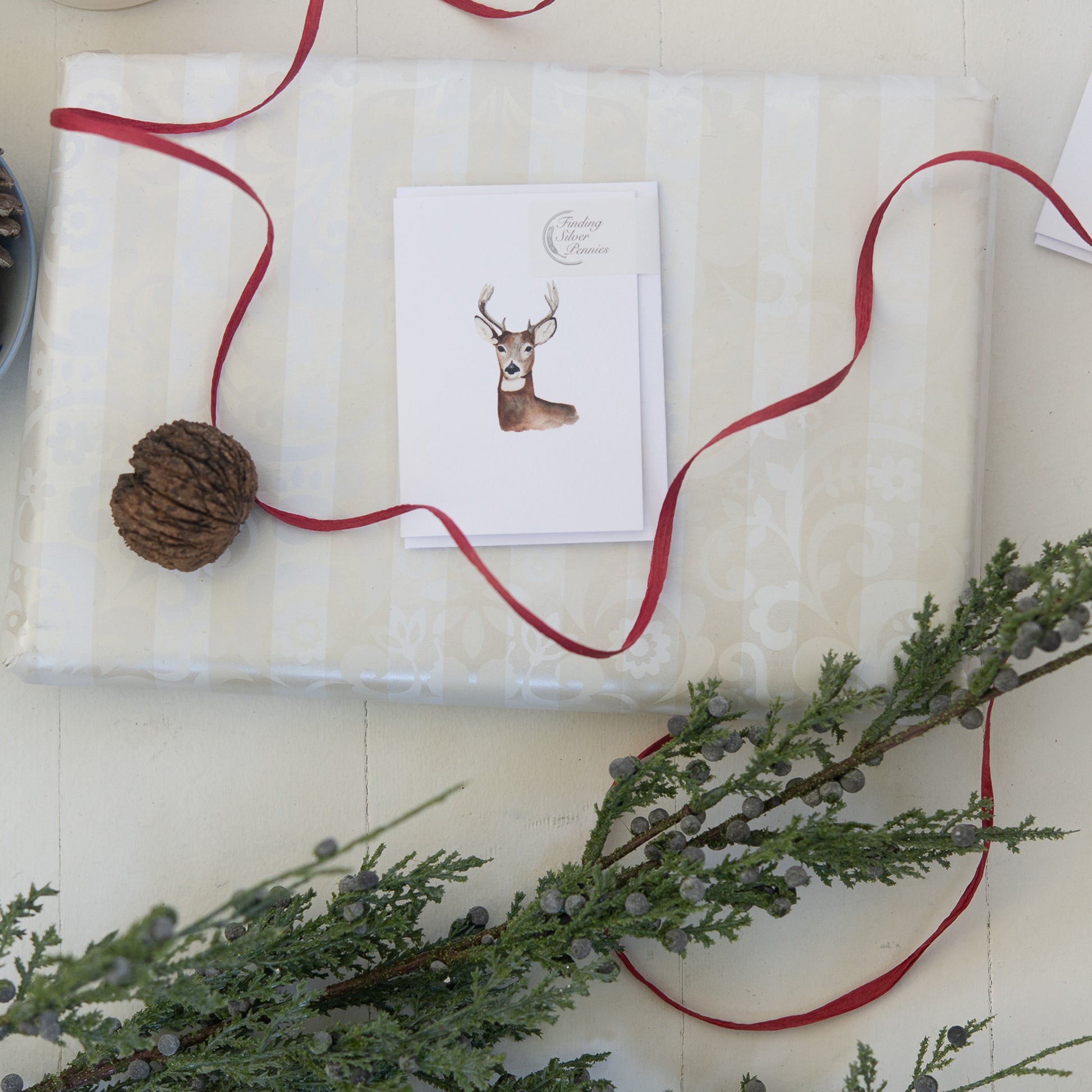 Gift wrapped in white paper with a red ribbon, pine cone, and card featuring a deer illustration on a light background.