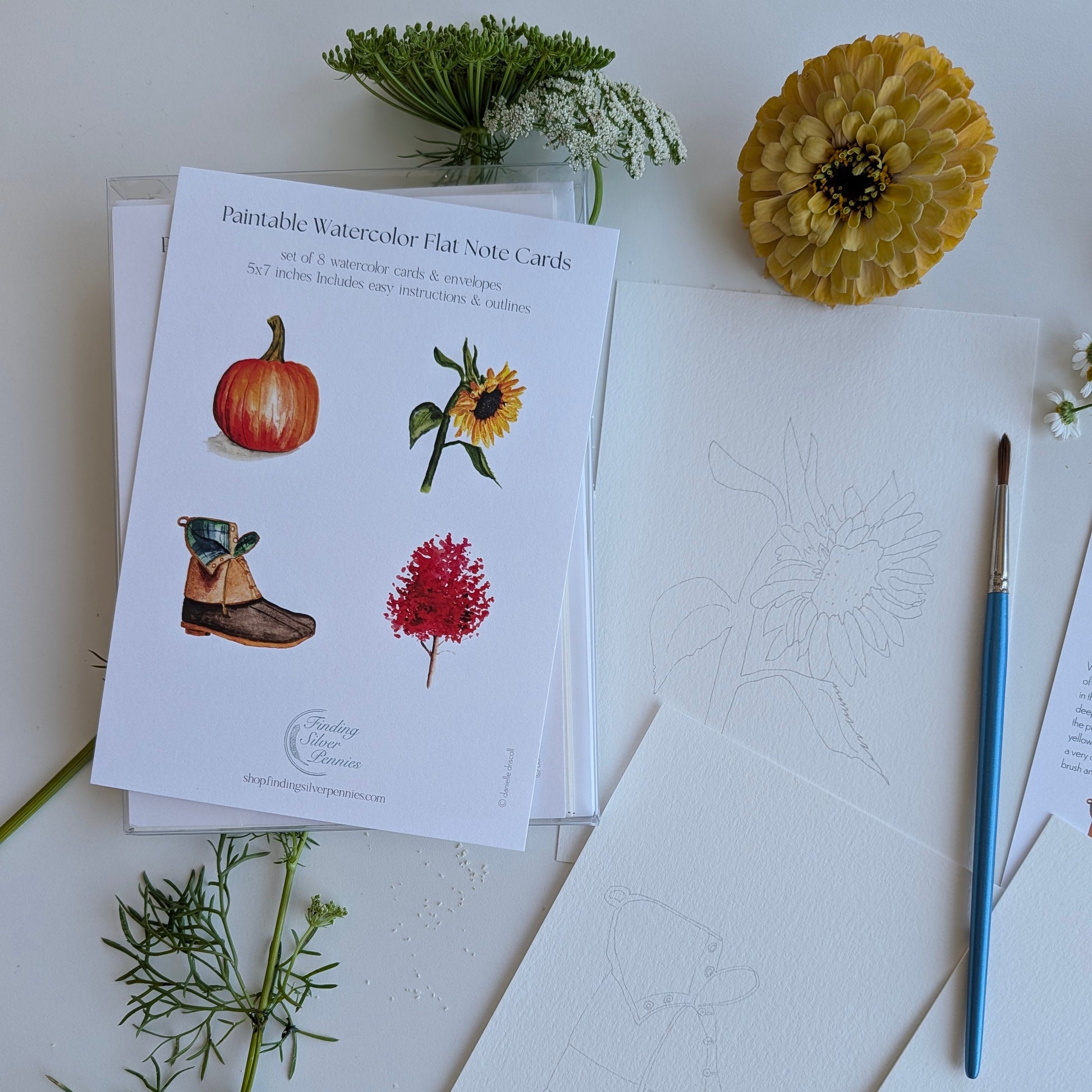 A styled flat-lay of watercolor note cards on a white surface, featuring printed cards with illustrations of a pumpkin, sunflower, boots, and red flowers, alongside blank textured cards, a paintbrush, and decorative flowers.