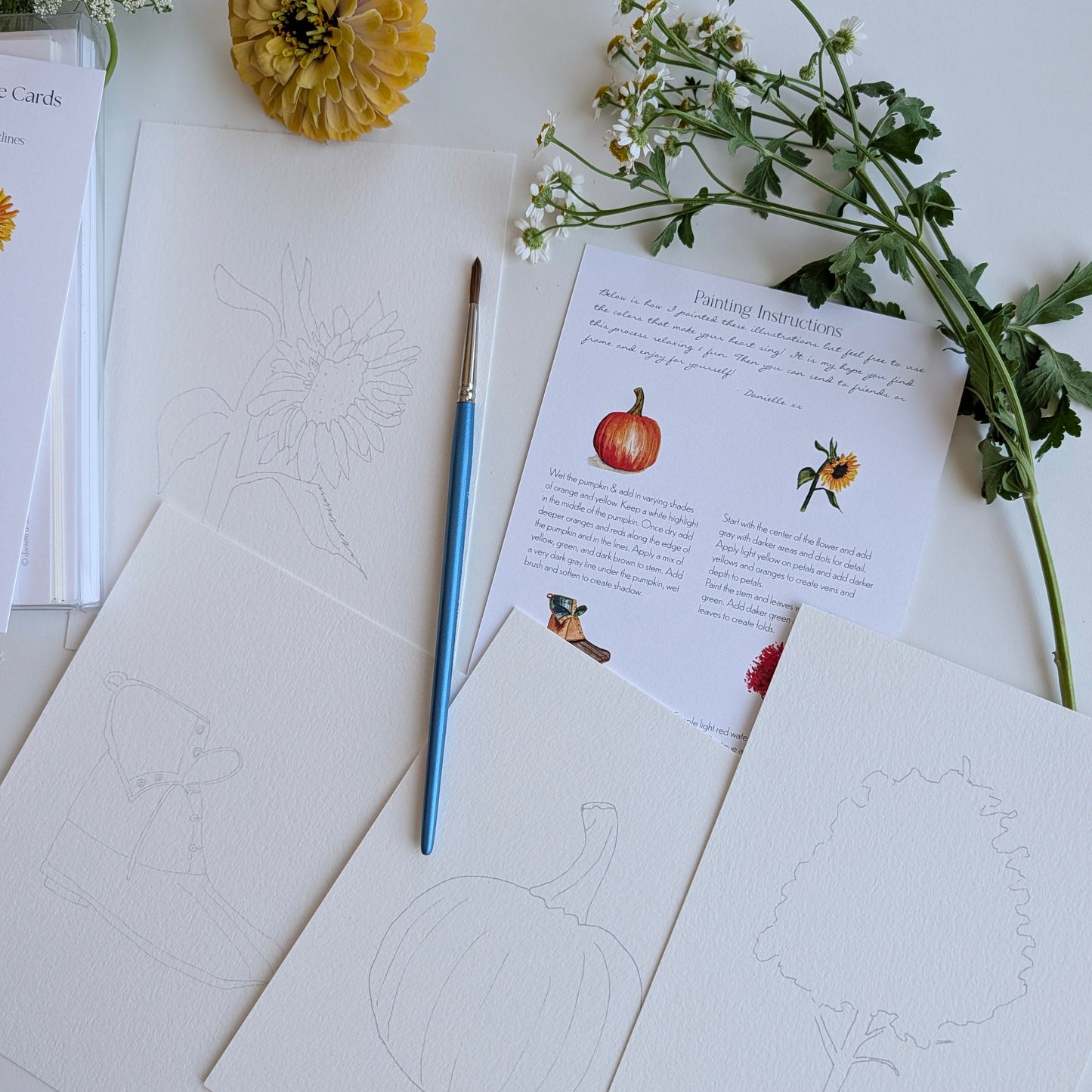 A styled flat-lay of a painting or coloring card set featuring autumn-themed line drawings including a pumpkin, tree, and flower, alongside a blue-handled paintbrush, a printed instruction sheet, and decorative flowers on a white background.