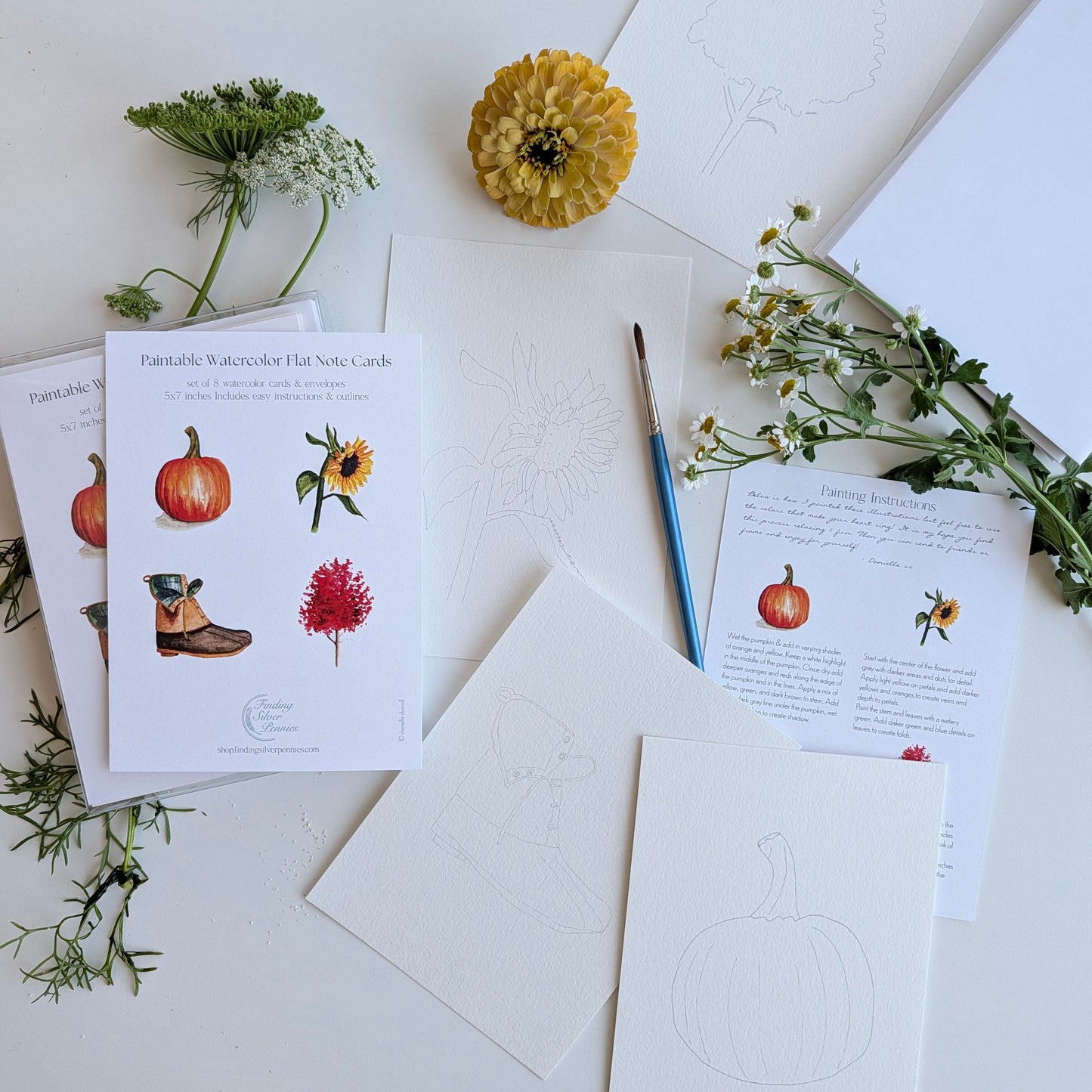 A styled flat-lay of printable watercolor hat box cards and art papers on a white surface, decorated with a yellow flower, green sprigs, and a paintbrush.