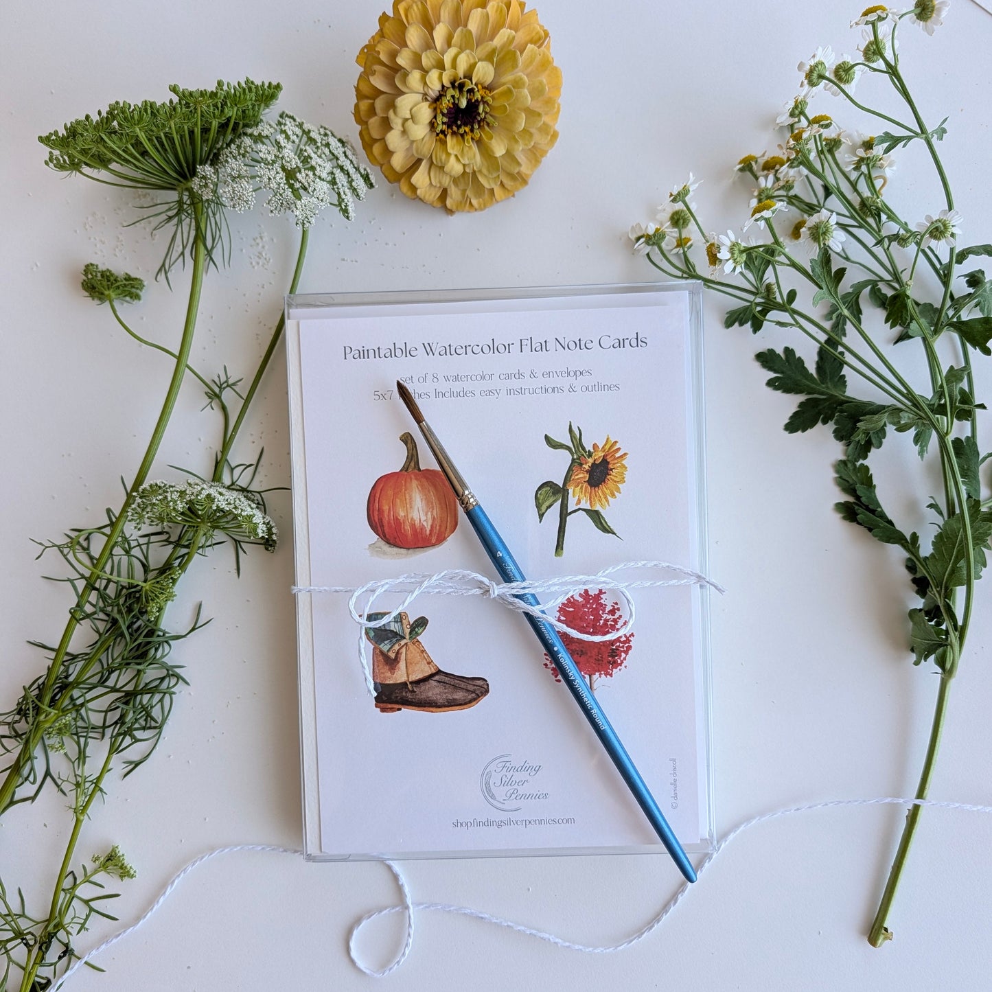 A flat note card set titled 'Paintable Watercolor Flat Note Cards' wrapped in white string, placed on a white surface with a blue brush, a yellow flower, and green botanical stems around it.