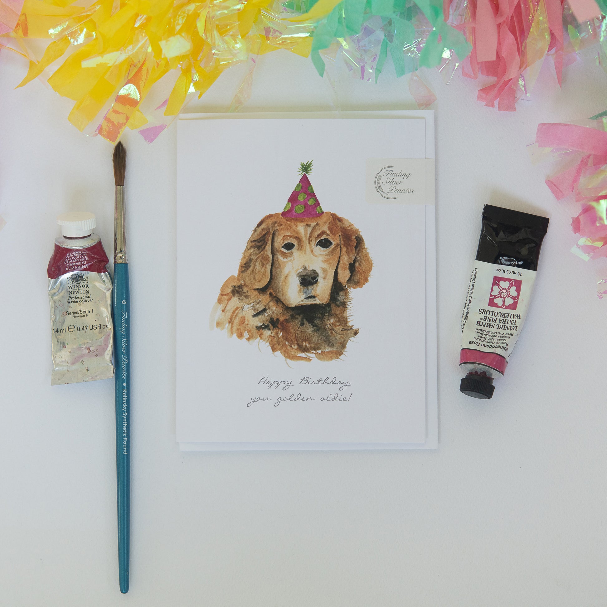 Painting of a dog wearing a party hat with art supplies and decorative ribbons on a white background.