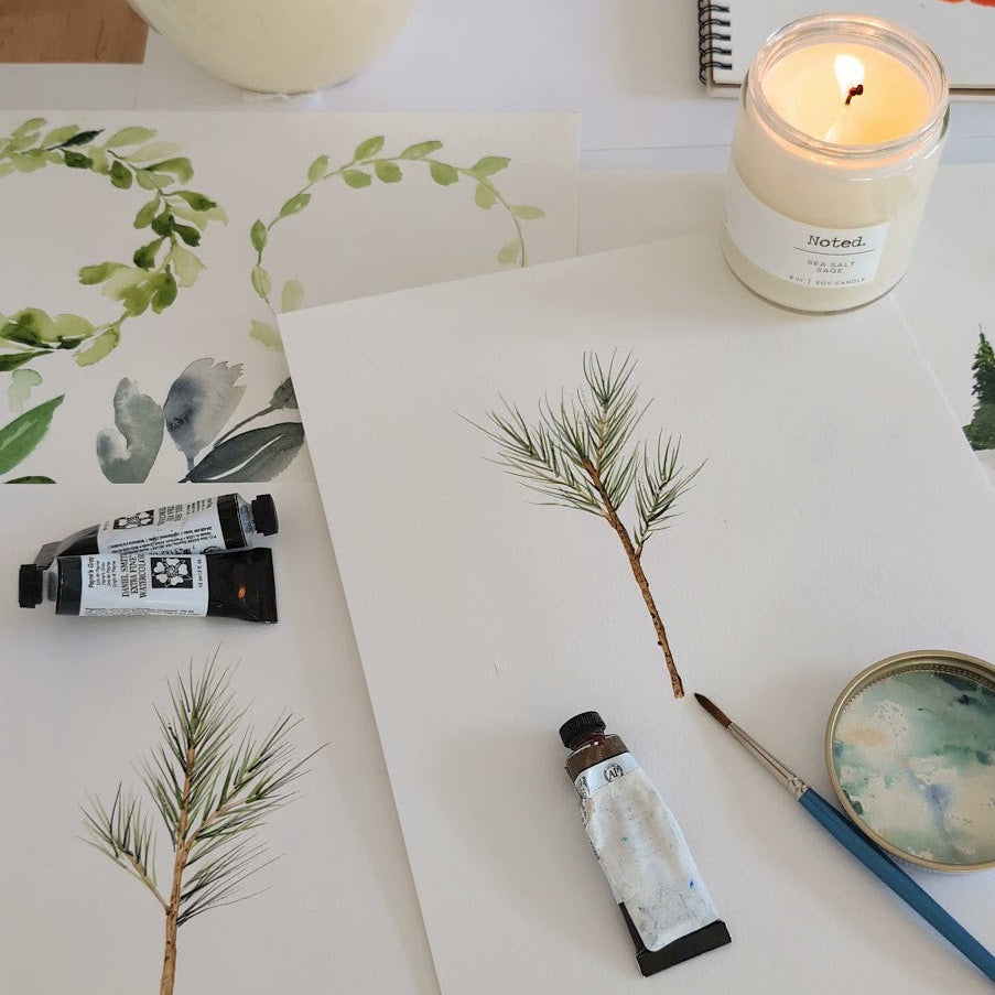An overhead flat-lay of an art studio setup featuring watercolor paints, a candle, a color wheel notebook, and painted pine branches on white paper.