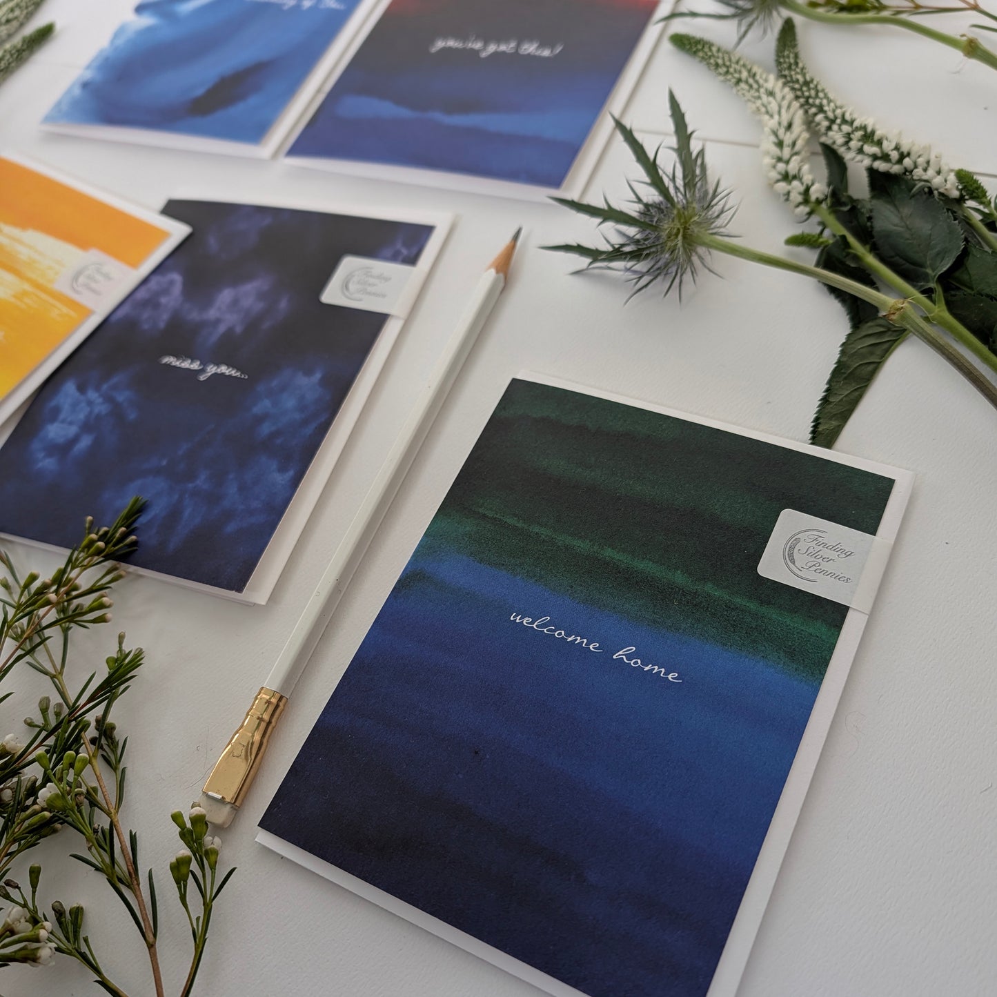 Greeting cards color washes and text on a white surface with greenery.