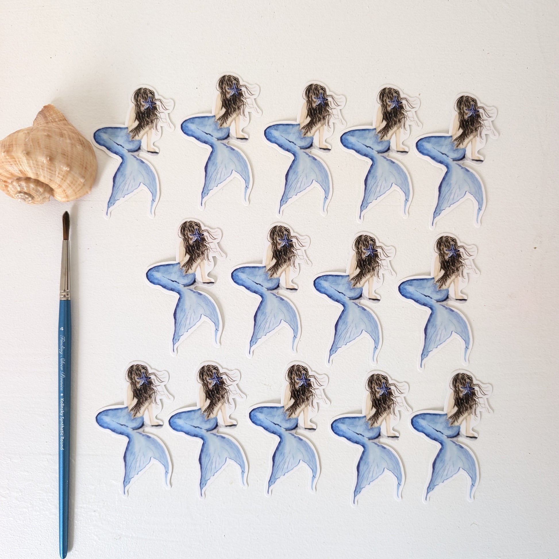 Multiple mermaid stickers with blue tails on a white background, accompanied by a paintbrush and shell.