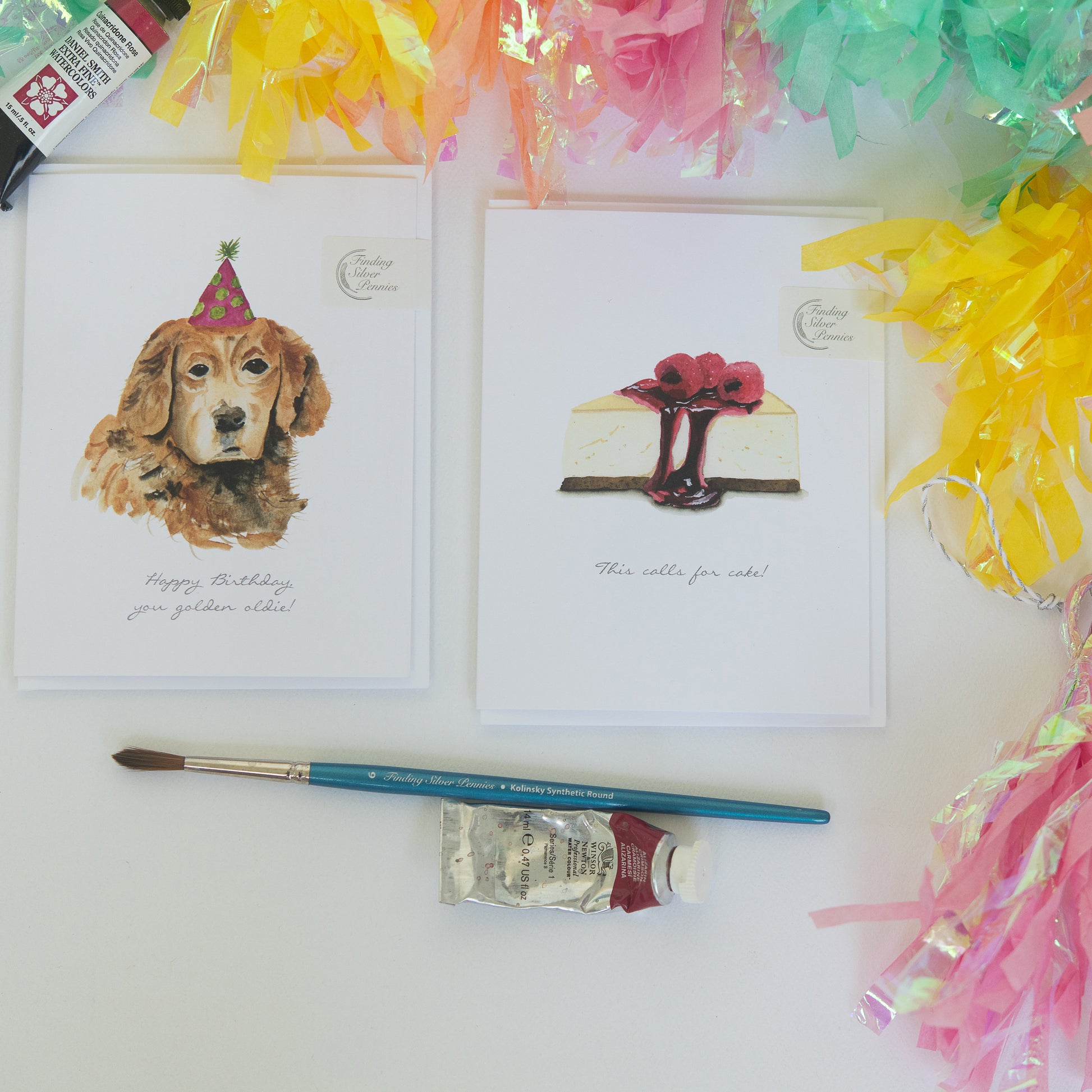 Two greeting cards with a dog and cake illustration, a paintbrush, and paint bottle on a white surface with colorful pom-poms.