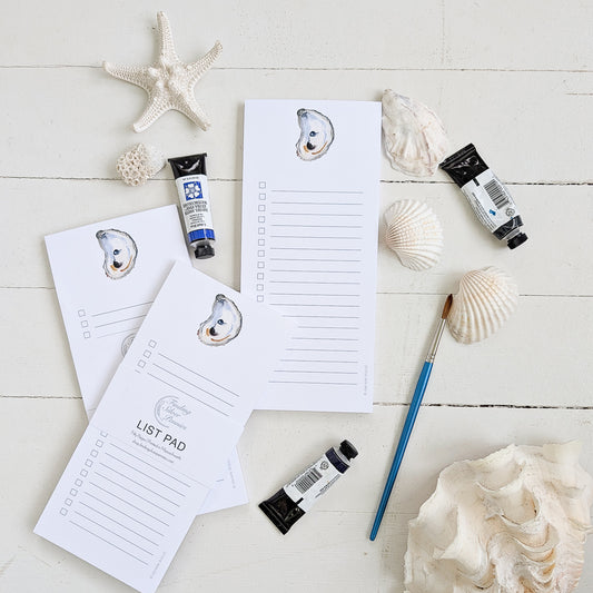 Note pads with shell designs, paint tubes, and a brush on a white wooden surface.