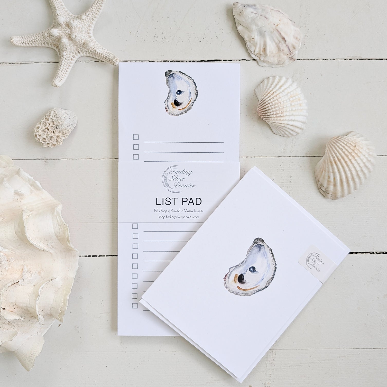 Two list pads with oyster designs on a wooden surface with seashells.