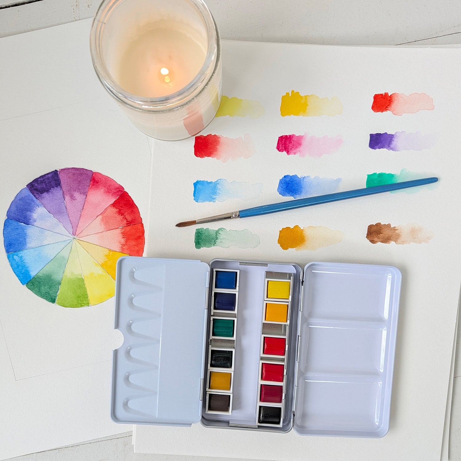Watercolor paint set with a color wheel, brush, and swatches on a white surface with a candle.