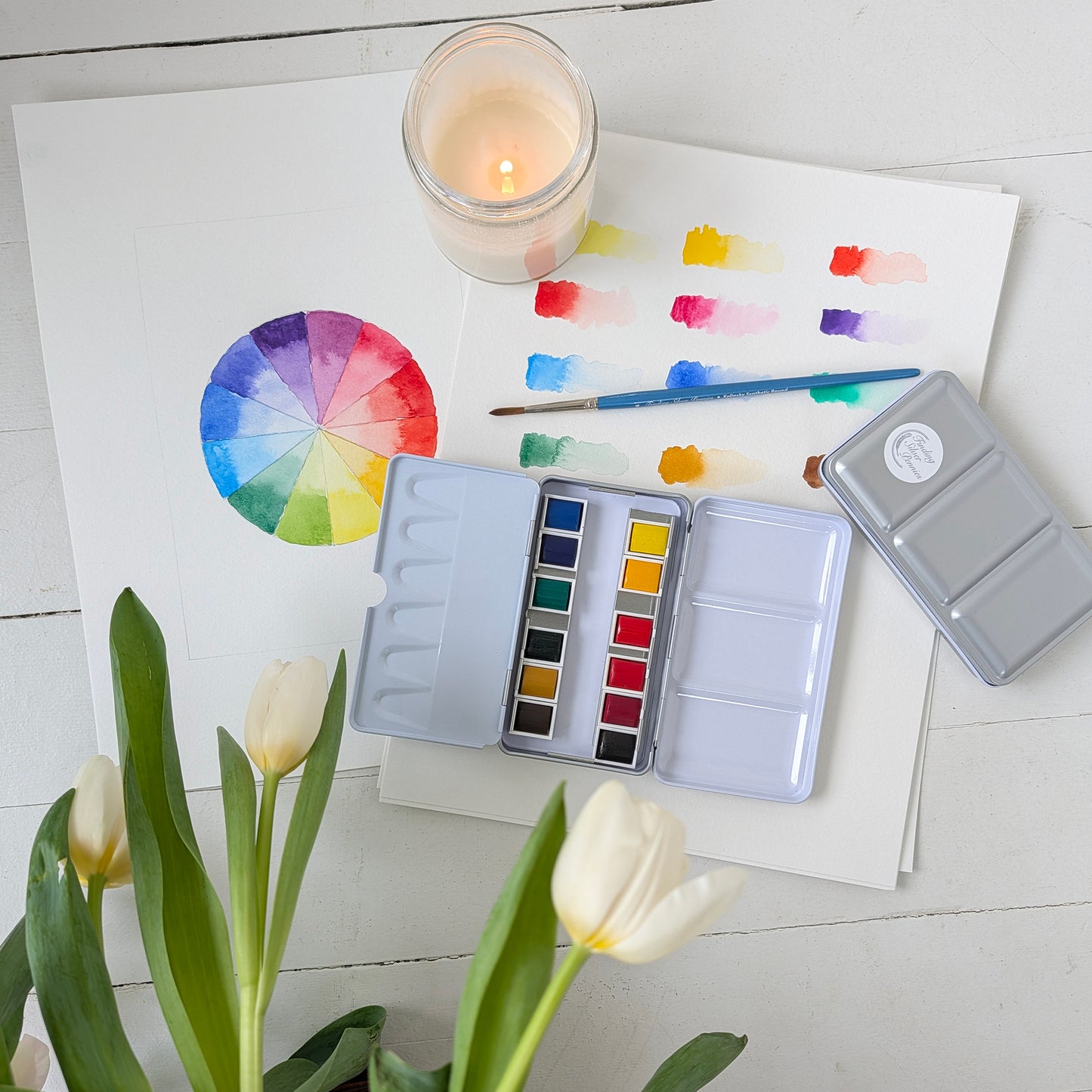 Artistic setup with watercolor paints, a color wheel, and tulips on a white surface.