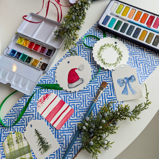 A styled flat-lay of watercolor painting supplies on a white table, including a watercolor palette, a paintbrush, greenery, and several decorated paper tags with painted holiday designs like a Santa hat, a wreath, and a tree.