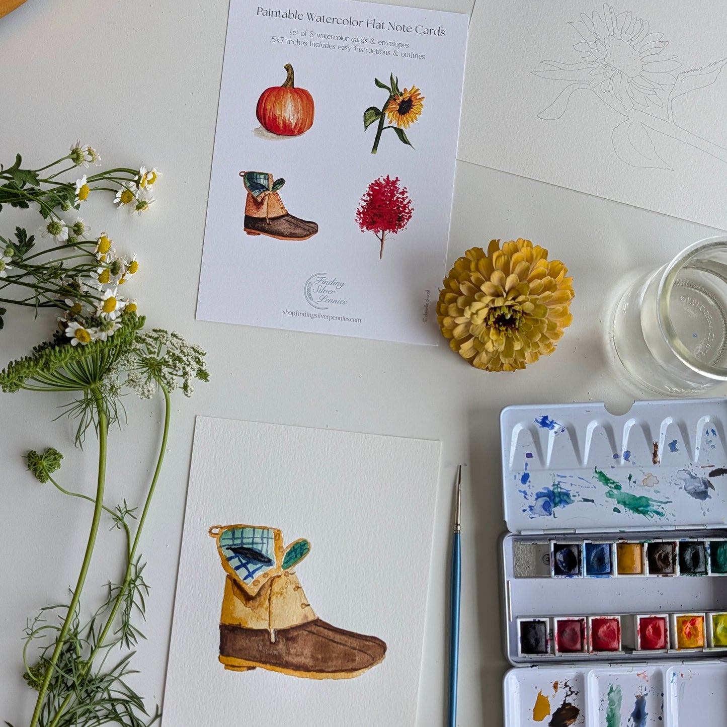 A styled flat-lay of watercolor art cards and painting supplies on a white surface. Two printed watercolor hair note cards are visible, one with illustrations of a pumpkin, sunflower, boot, and red flower, and another card shows a partially painted brown boot.