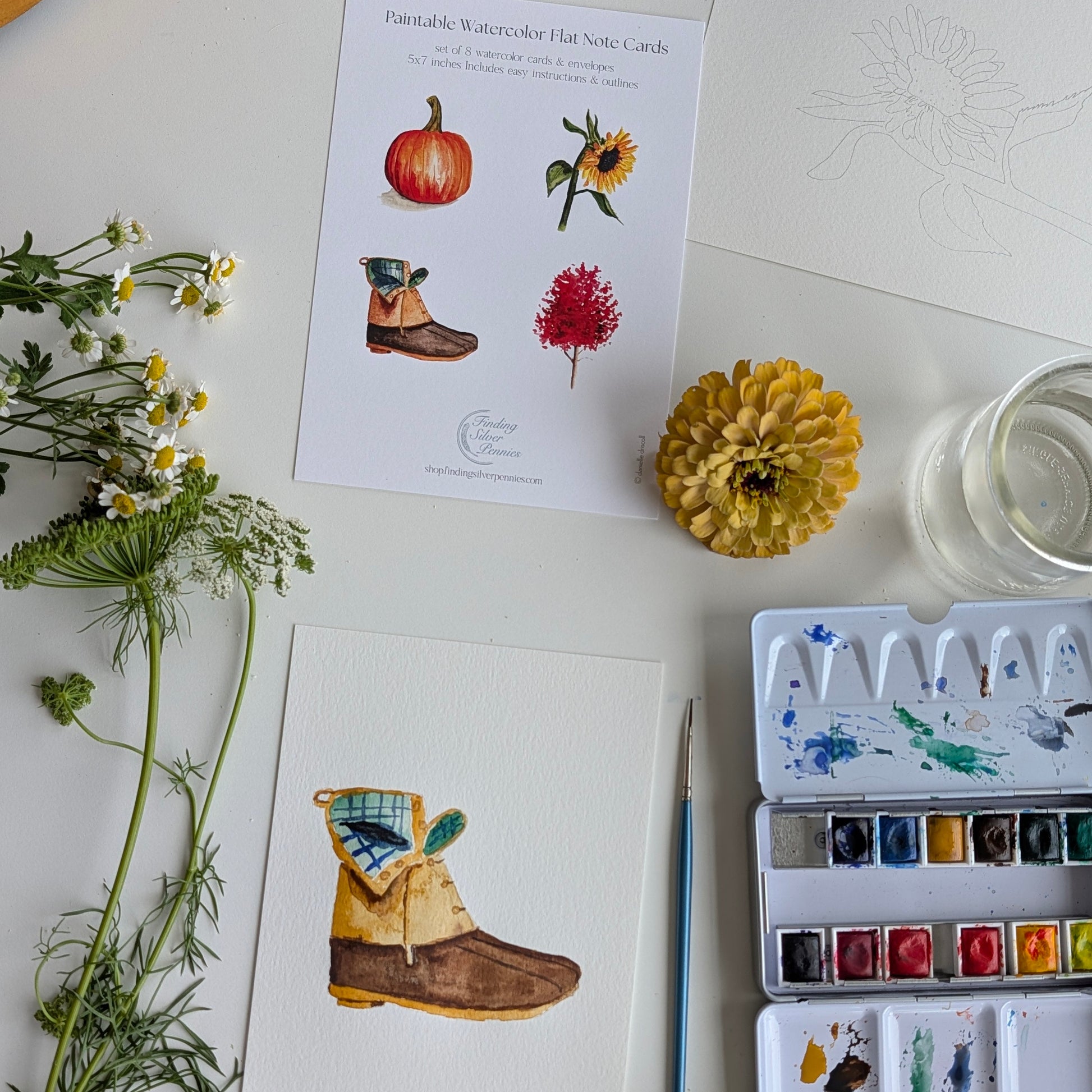 A styled flat-lay of watercolor art cards and painting supplies on a white surface. Two printed watercolor hair note cards are visible, one with illustrations of a pumpkin, sunflower, boot, and red flower, and another card shows a partially painted brown boot.