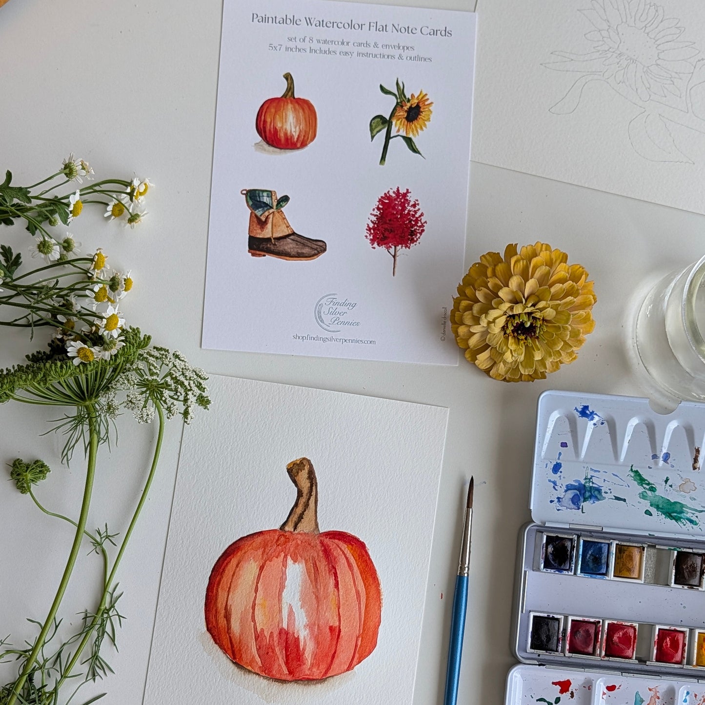 A styled flat-lay of watercolor flat note cards and painting supplies on a white surface. A printed card at the top shows watercolor illustrations including a pumpkin, sunflower, boot, and red flower.