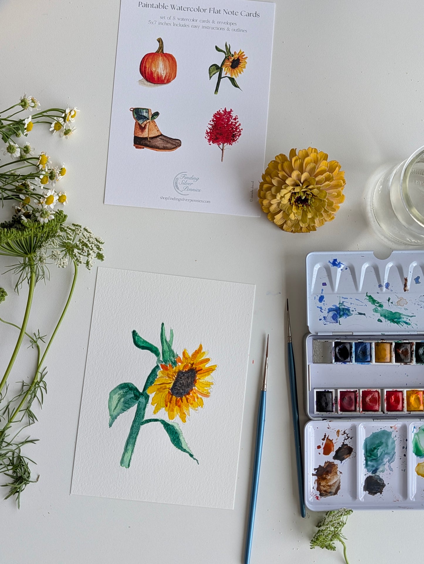A styled flat-lay of watercolor art supplies on a white surface, including a printed watercolor flower card, a painted sunflower note card, a paint palette with mixed colors, paintbrushes, flowers, and a glass of water.
