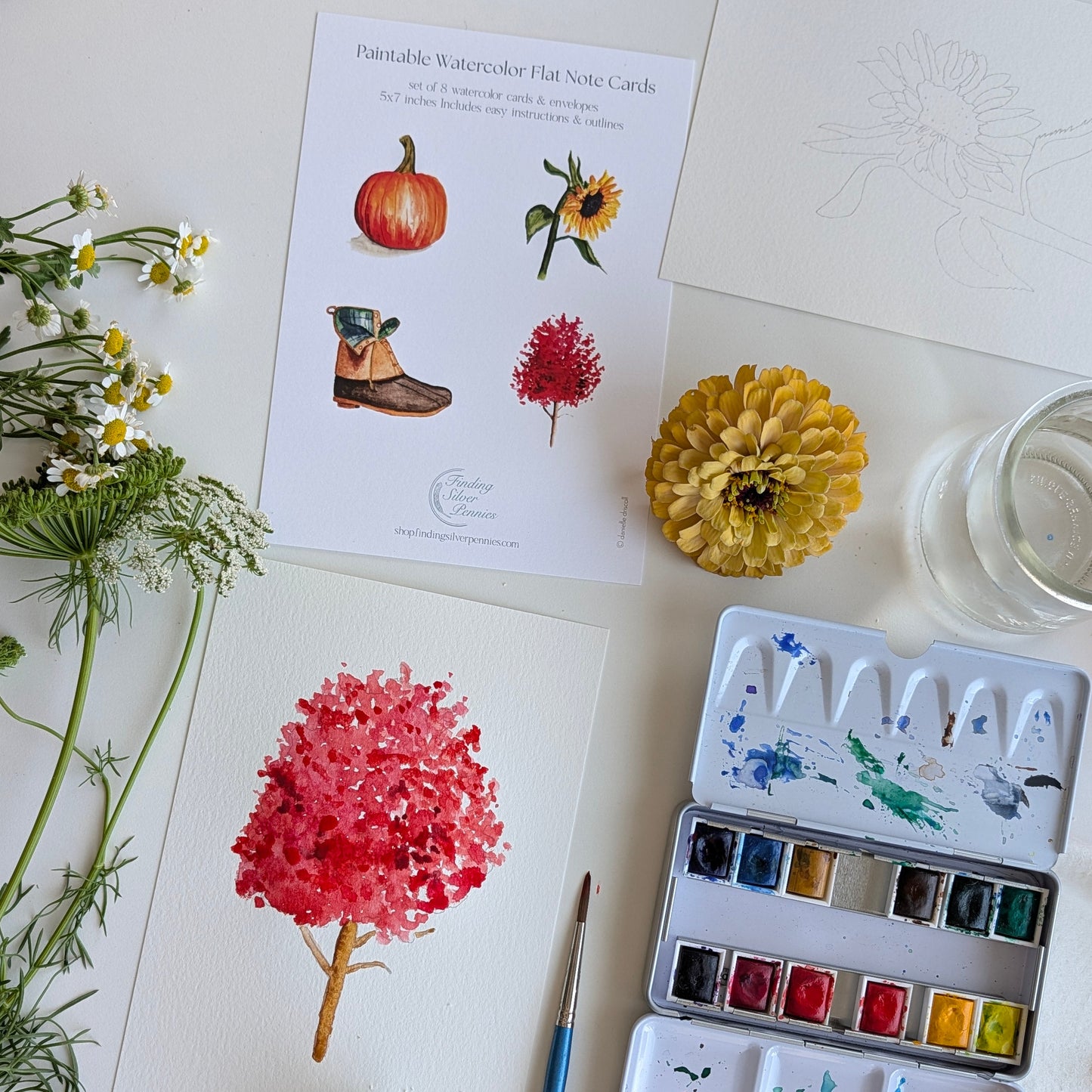 A styled flat-lay of watercolor painting supplies on a white surface, including a watercolor paint card sheet, a watercolor palette with mixed paints, a paintbrush, a yellow flower, white daisies, and a glass of water.
