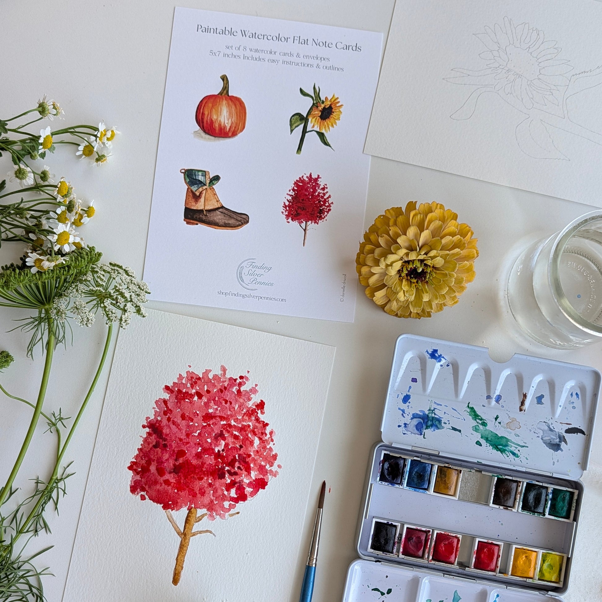 A styled flat-lay of watercolor painting supplies on a white surface, including a watercolor paint card sheet, a watercolor palette with mixed paints, a paintbrush, a yellow flower, white daisies, and a glass of water.