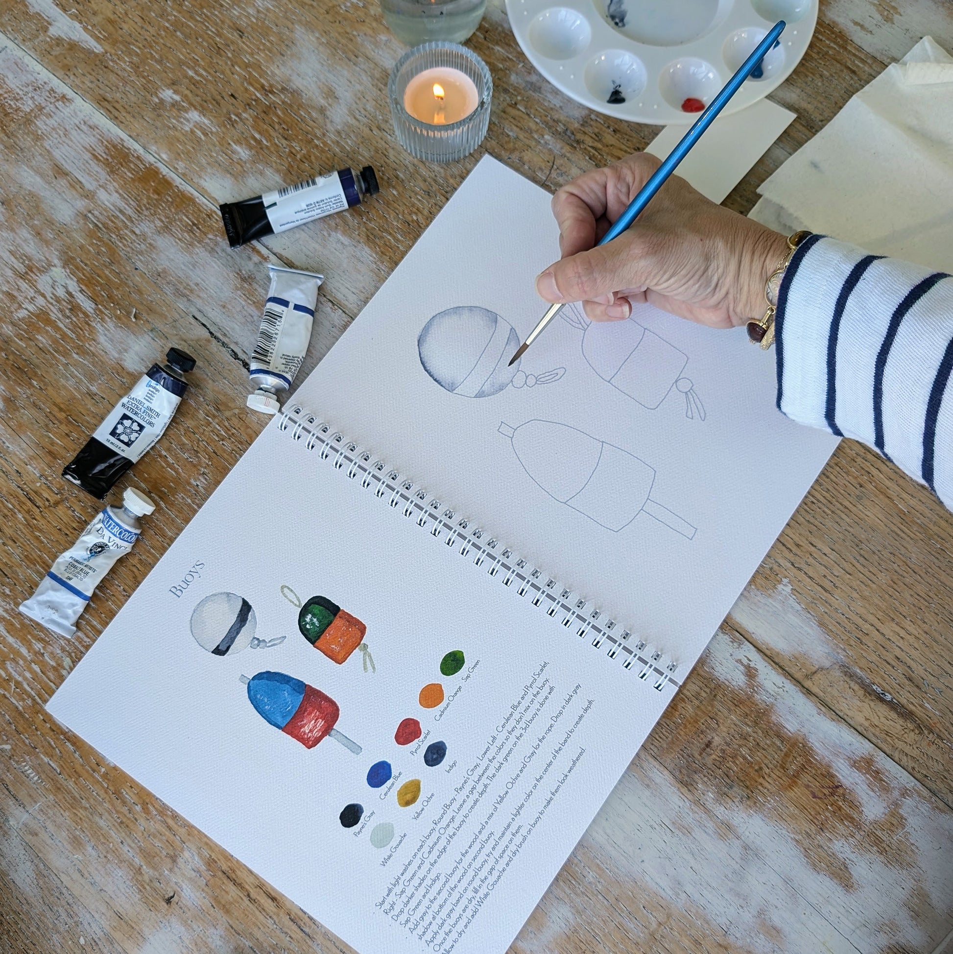 Person painting buoys in watercolor workbook surrounded by paint supplies