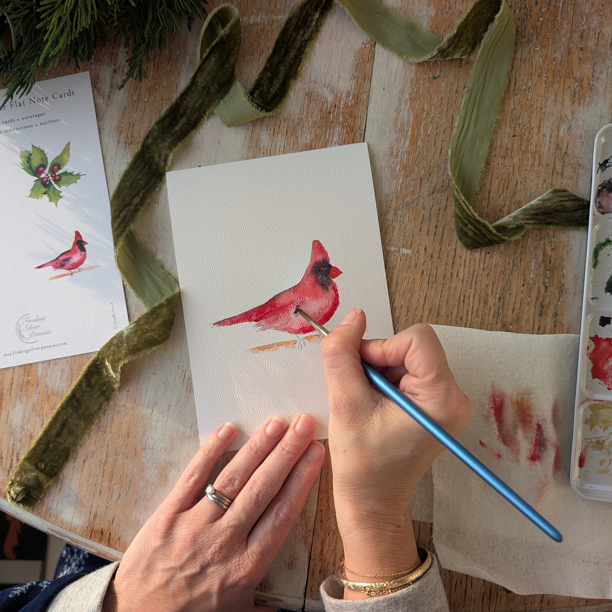 Person painting a red cardinal on a piece of paper with watercolors, surrounded by art supplies on a wooden table.