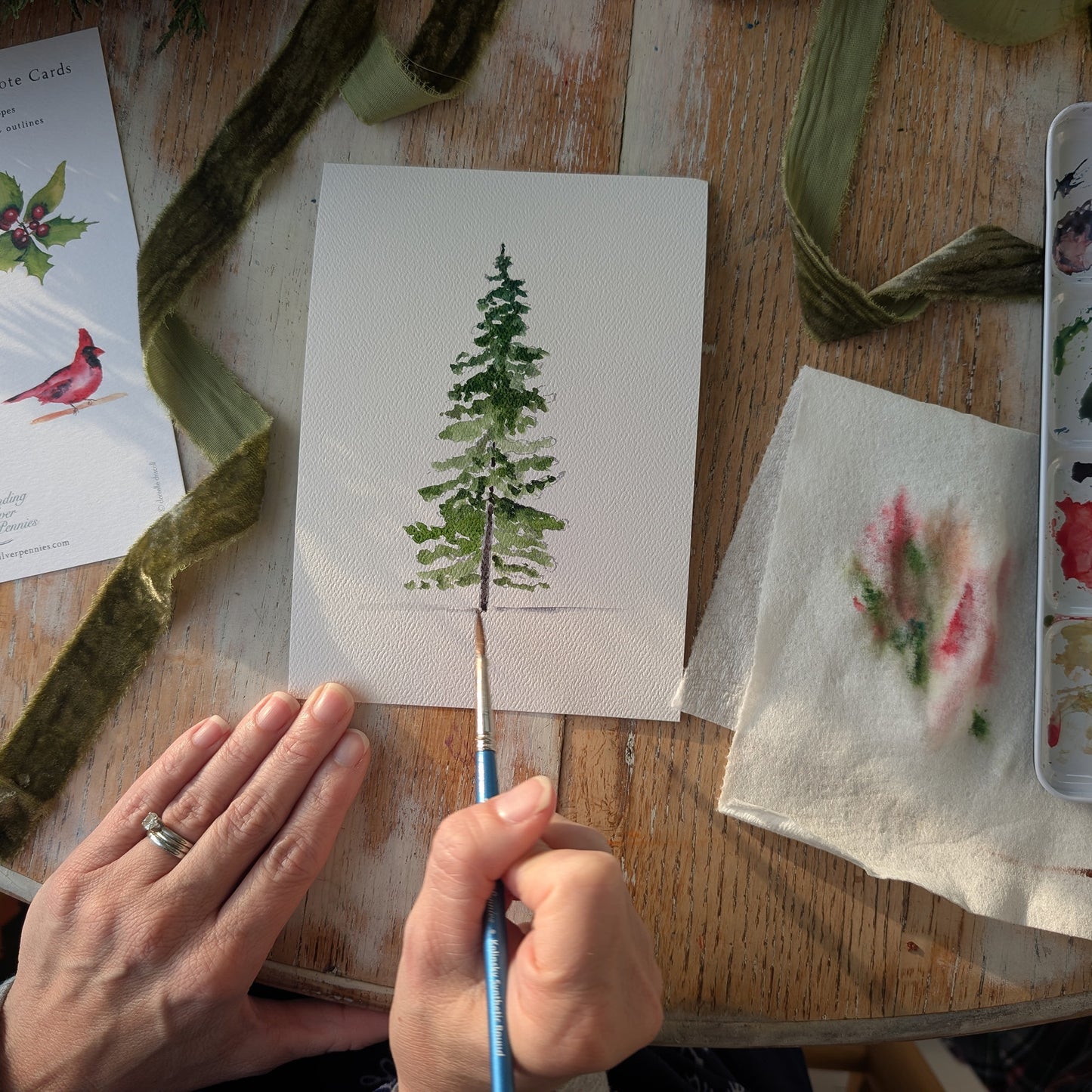 Person painting a green tree on a small canvas with watercolor paints and brushes on a wooden table.