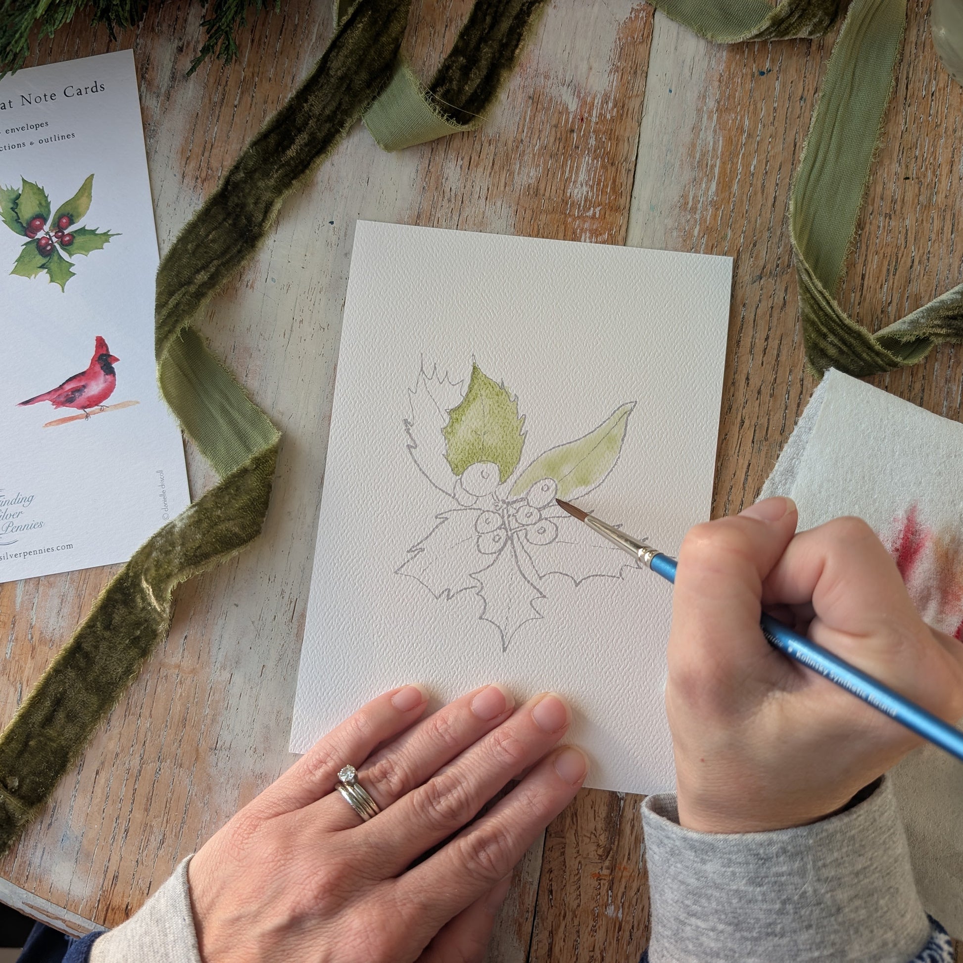 Person painting a leaf design on a card with a brush, surrounded by green ribbons and a card with a bird illustration.