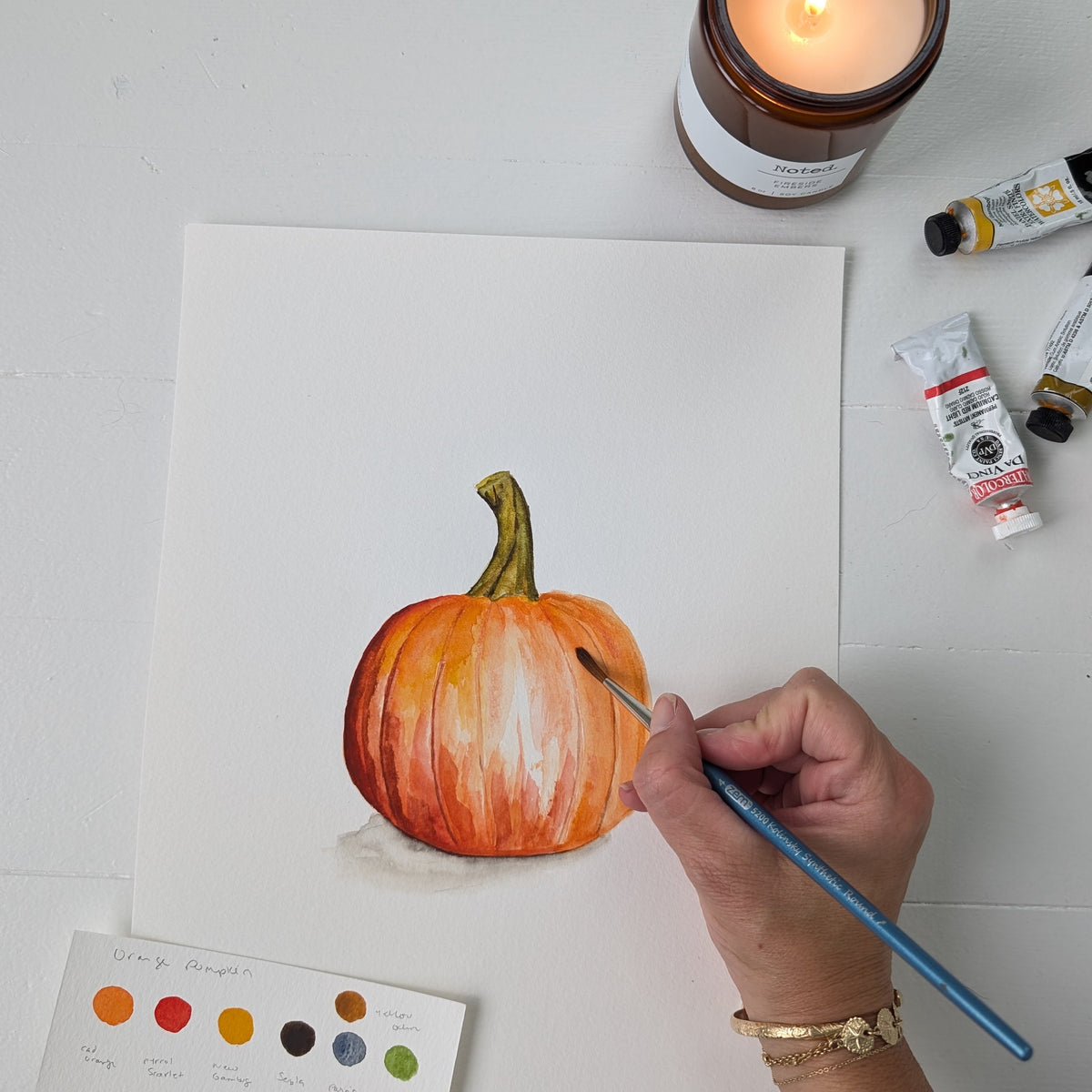 A hand holds a paintbrush over a watercolor painting of a pumpkin on white paper, arranged on a white tabletop with a lit candle, paint tubes, and a color card nearby.
