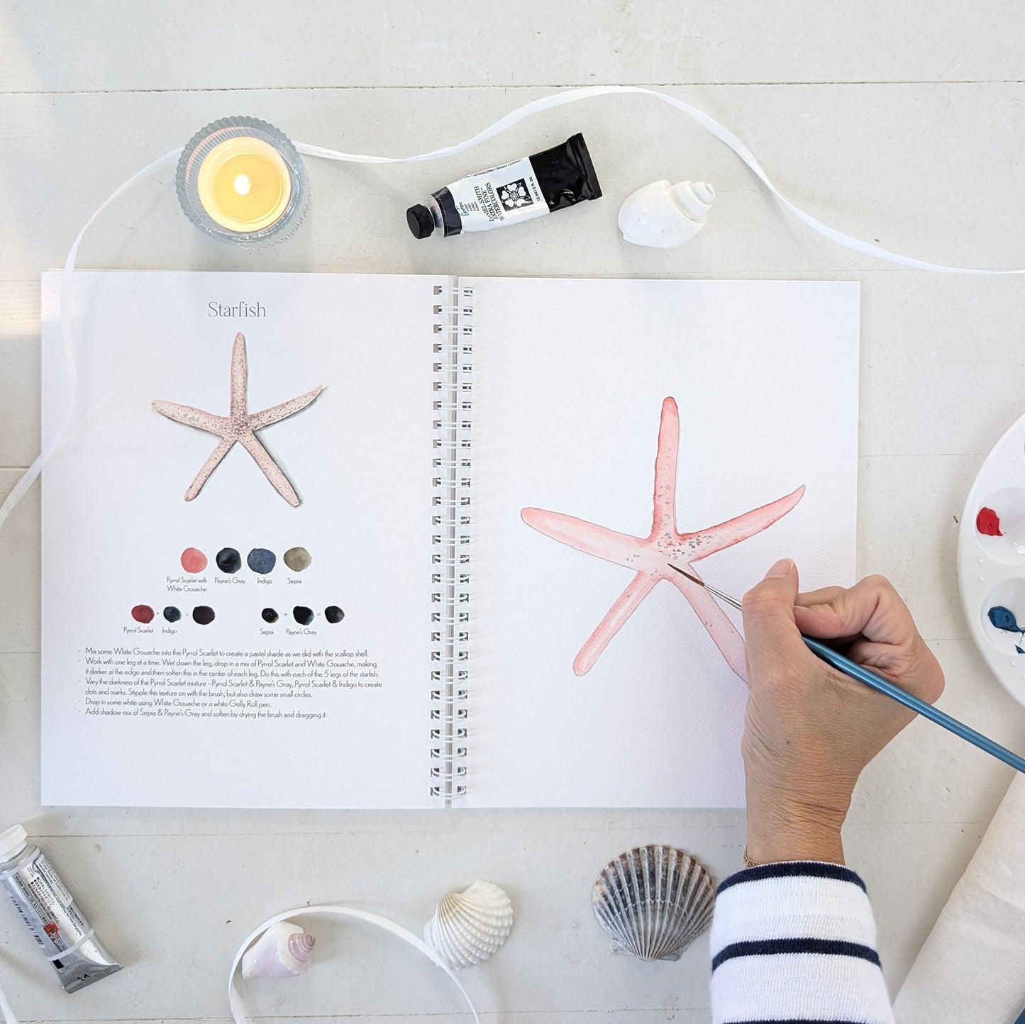 Person painting a starfish in a watercolor workbook with art supplies on a white surface