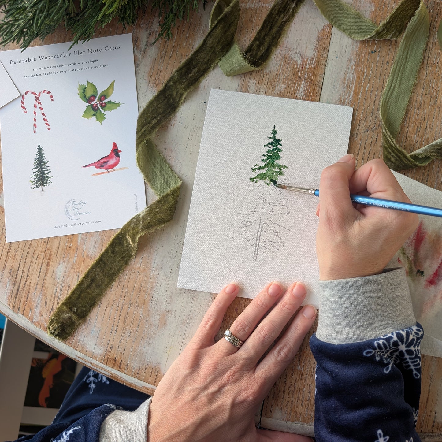Person painting a tree on a piece of paper with green yarn and watercolor cards in the background.