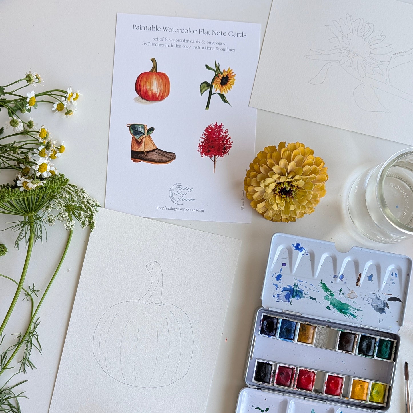A styled flat-lay of a watercolor painting set on a white surface, including a watercolor paint palette with mixed paint colors, a printed card titled 'Paintable Watercolor Flat Note Cards,' a blank pumpkin sketch, flowers, and a glass of water.