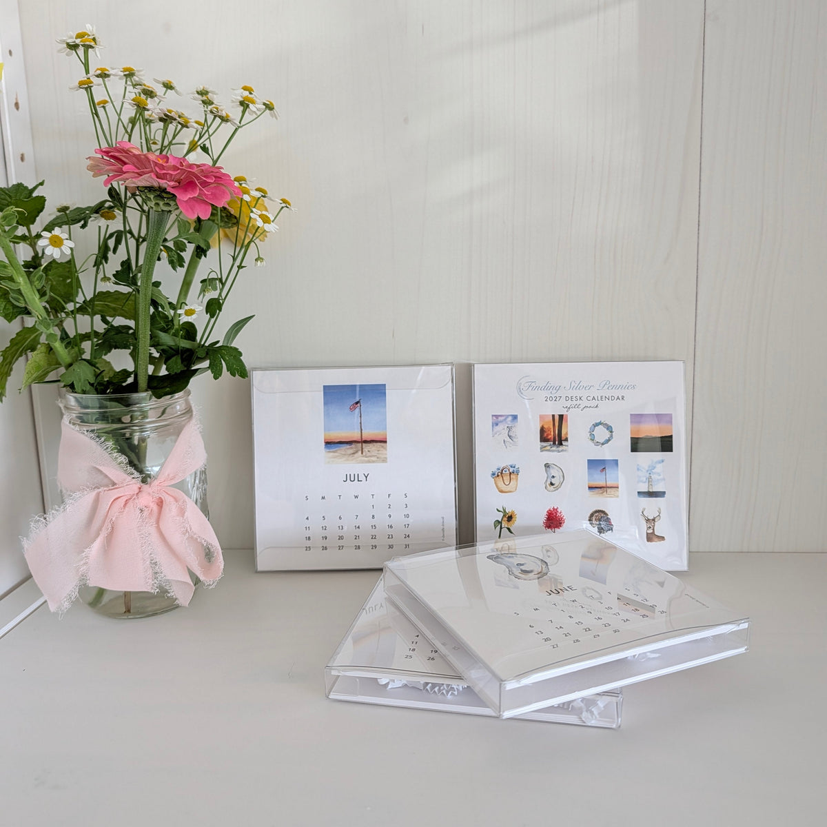 A styled tabletop scene with a glass jar of pink and yellow flowers tied with a pink ribbon, two small printed cards or calendars, and a stack of clear plastic-wrapped items on a white surface against a white paneled background.