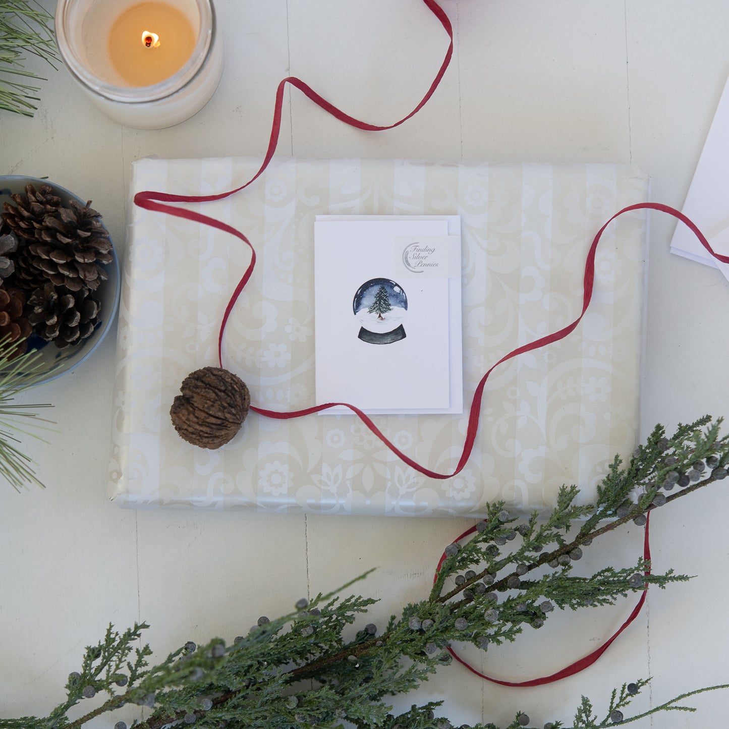 Gift wrapped in white paper with a red ribbon, surrounded by pine cones and greenery on a light surface.